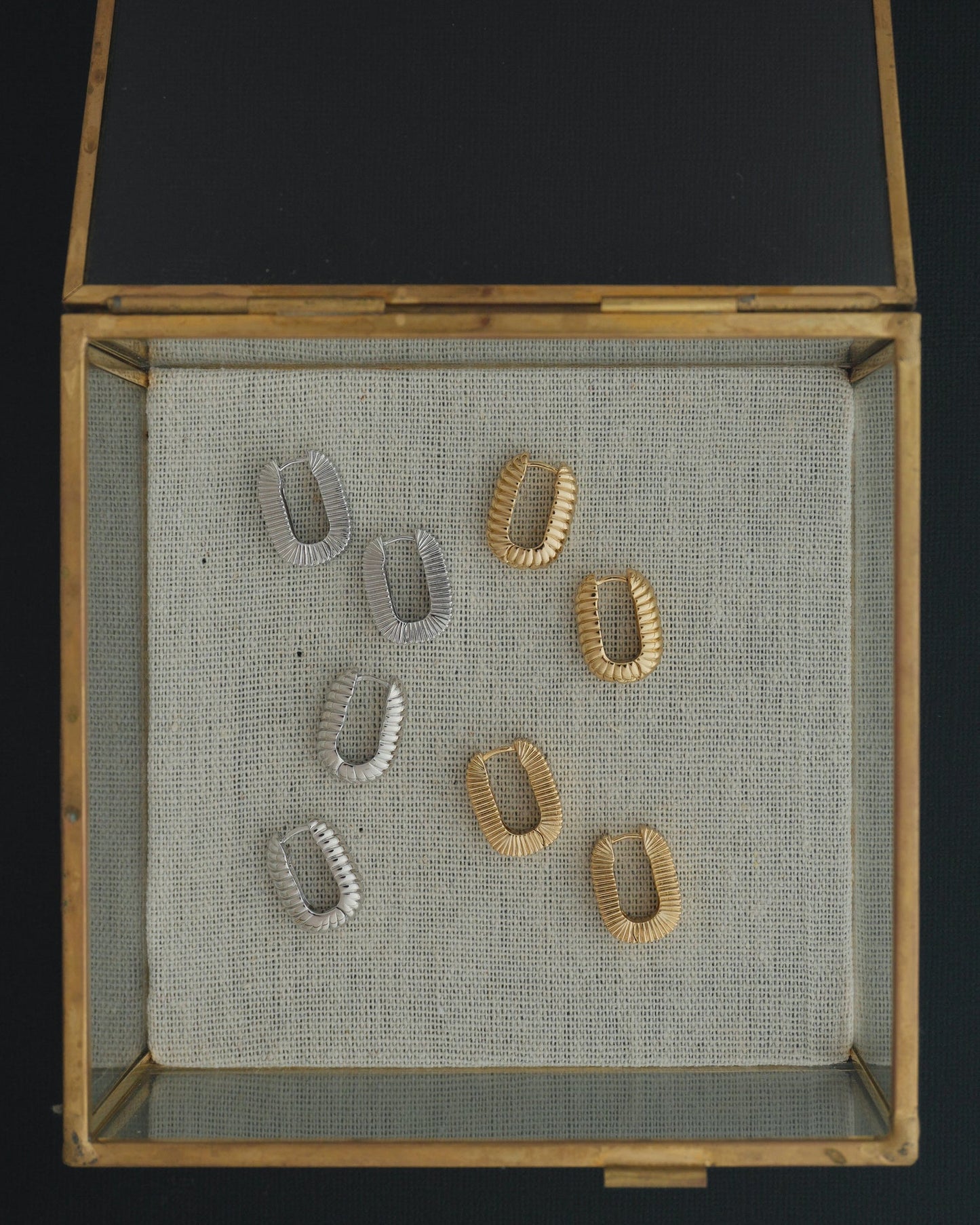 A glass and brass display box features four pairs of Mod + Jo Elly French Rope Hoops—including two shimmering gold and two silver pairs—beautifully arranged on a textured beige fabric background. These elegant earrings combine timeless style with lightweight versatility, making them perfect for those who love the classic look of hoop earrings.