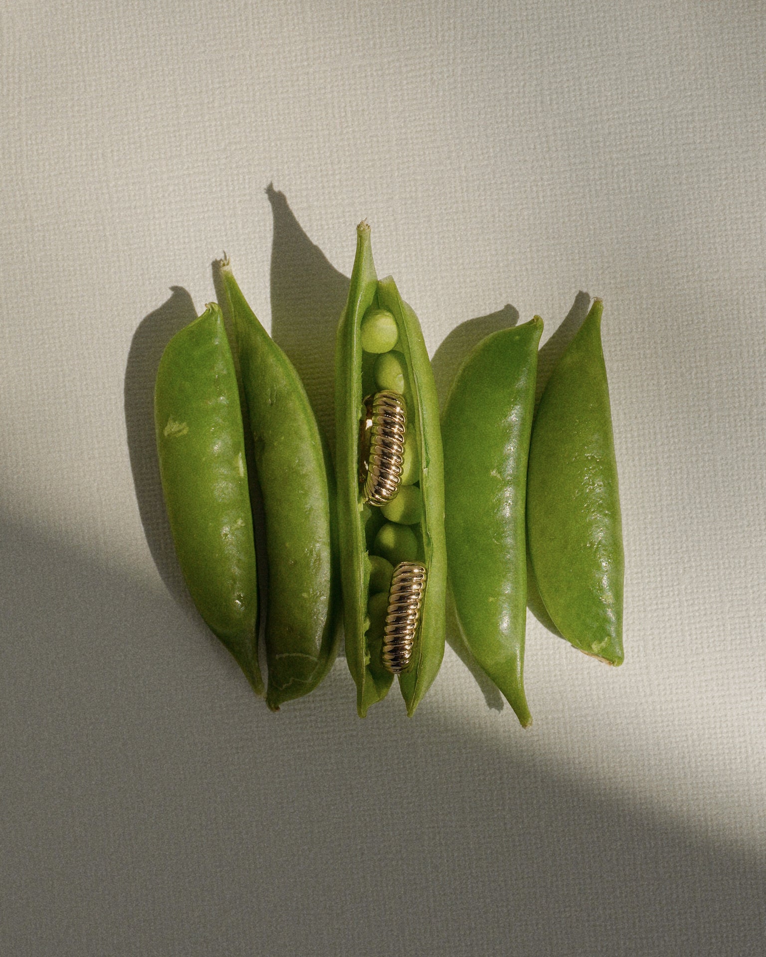 Four pea pods rest on a textured surface; the middle pod is open, revealing two golden screw coils nestled among the peas—an artful fusion of nature and design reminiscent of Mod + Jo’s Elly French Rope Hoops.