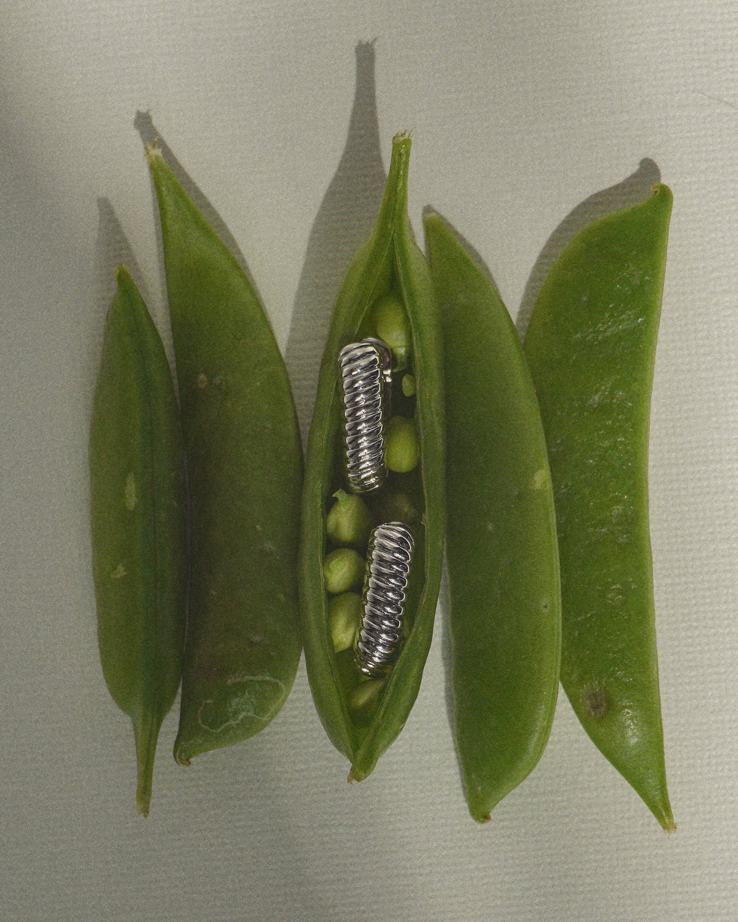 Four green pea pods are displayed, with one pod open to reveal two metal springs nestled among the peas—an allusion to the surprising design of the Elly French Rope Hoops by Mod + Jo. The image artfully blends organic and metallic elements against a neutral backdrop.