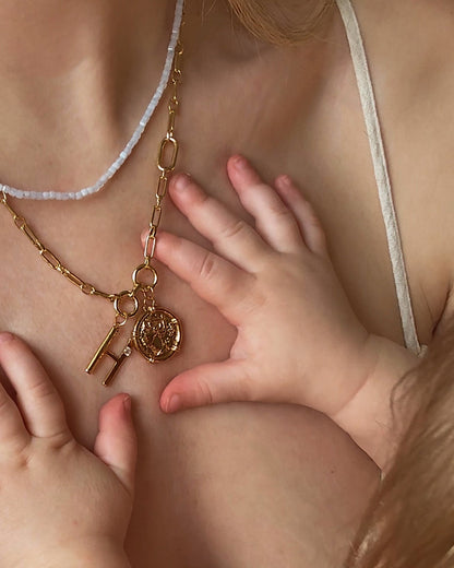 A close-up of a baby’s hands holding Mod + Jo’s Eloise Letter Charm necklace and a gold round pendant, both worn by an adult with bare skin and a white strap.