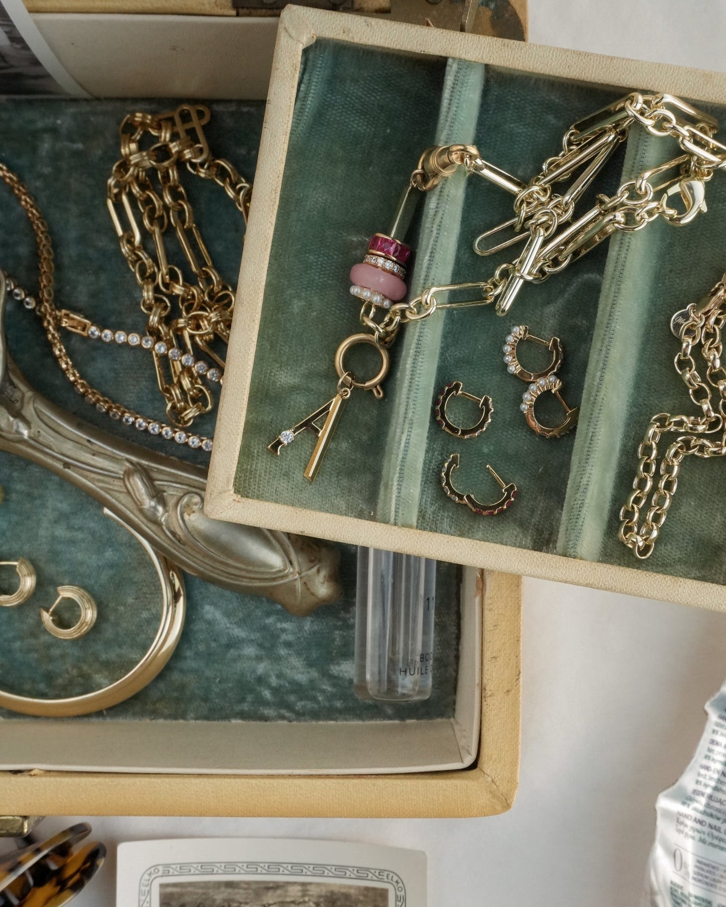An open jewelry box reveals the Mod + Jo Liam Space Bar Chain Necklace arranged on green velvet lining, alongside hoop earrings, a charm with beads, a gold bangle, and a small clear vial.