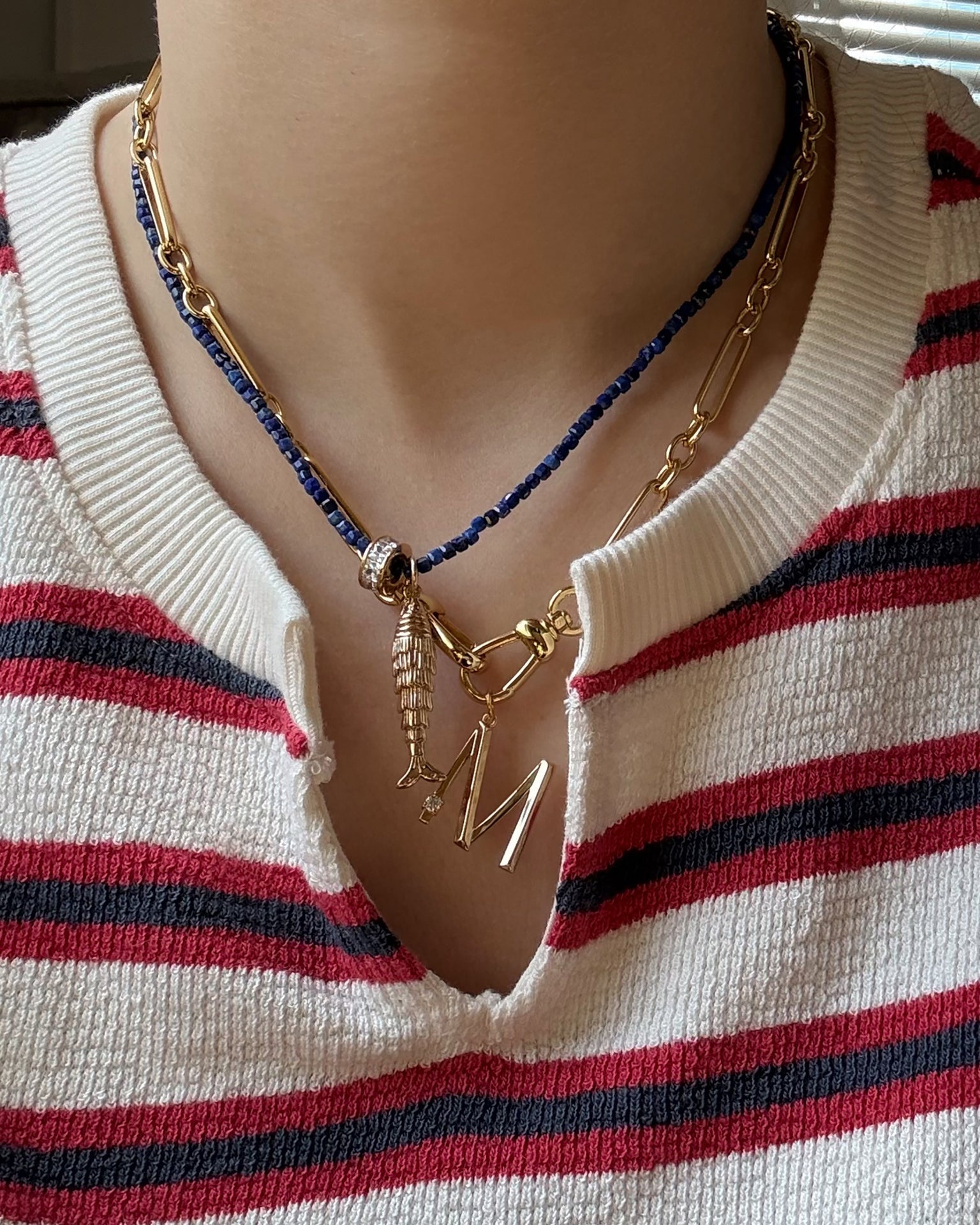A person wears a white and red striped shirt with three layered necklaces: a gold chain, dark beaded strand, and Mod + Jo's Eloise Letter Charm—an 18k gold plated necklace with a fish charm and large gold initial M.