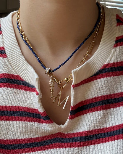 A person wears a white and red striped shirt with three layered necklaces: a gold chain, dark beaded strand, and Mod + Jo's Eloise Letter Charm—an 18k gold plated necklace with a fish charm and large gold initial M.