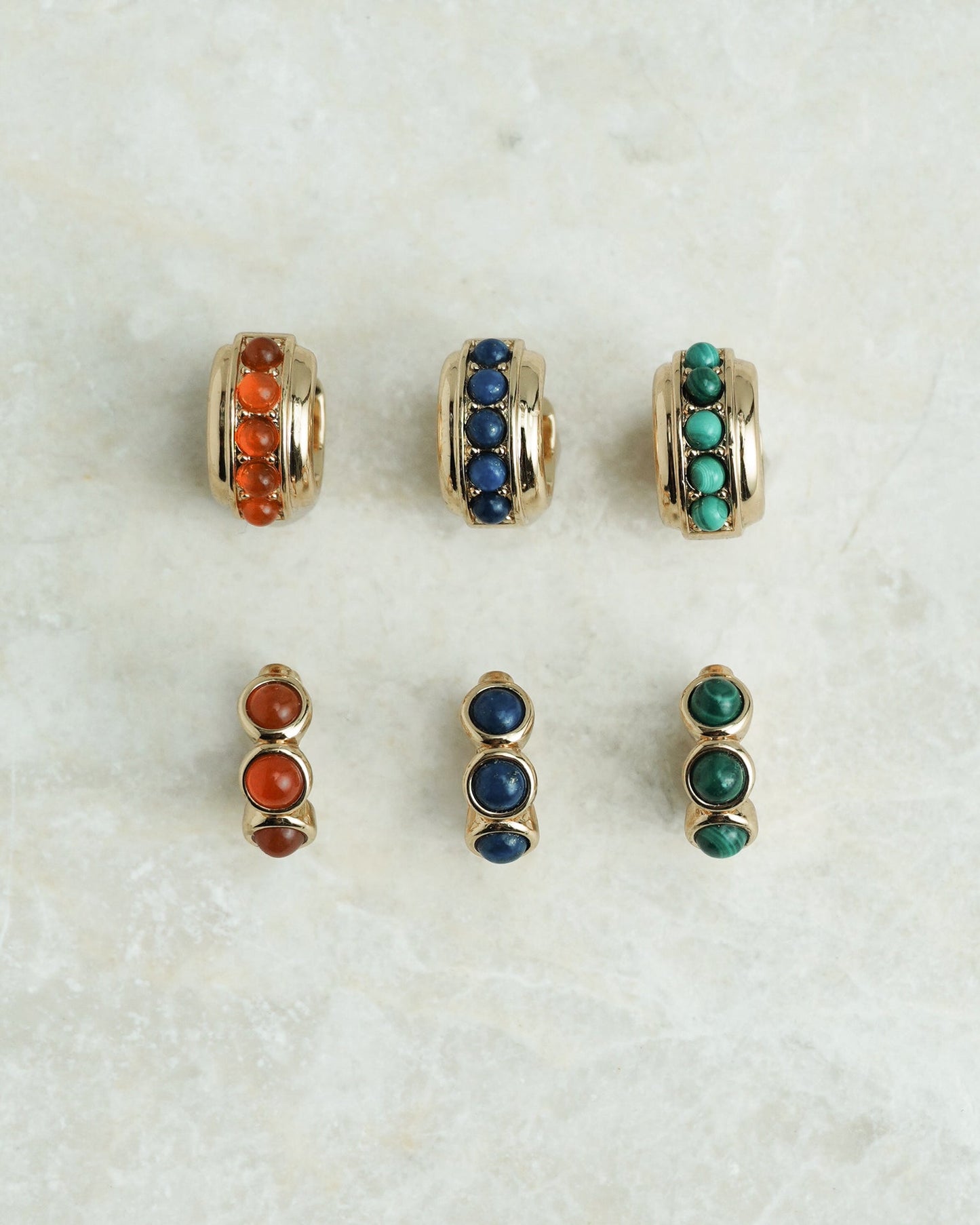 Six pairs of Mod + Jo Corynn Gemstone Huggies, plated in 18k gold with vibrant red, lapis lazuli blue, and green round stones, are arranged on a light marble surface in sets of two and three for each color.