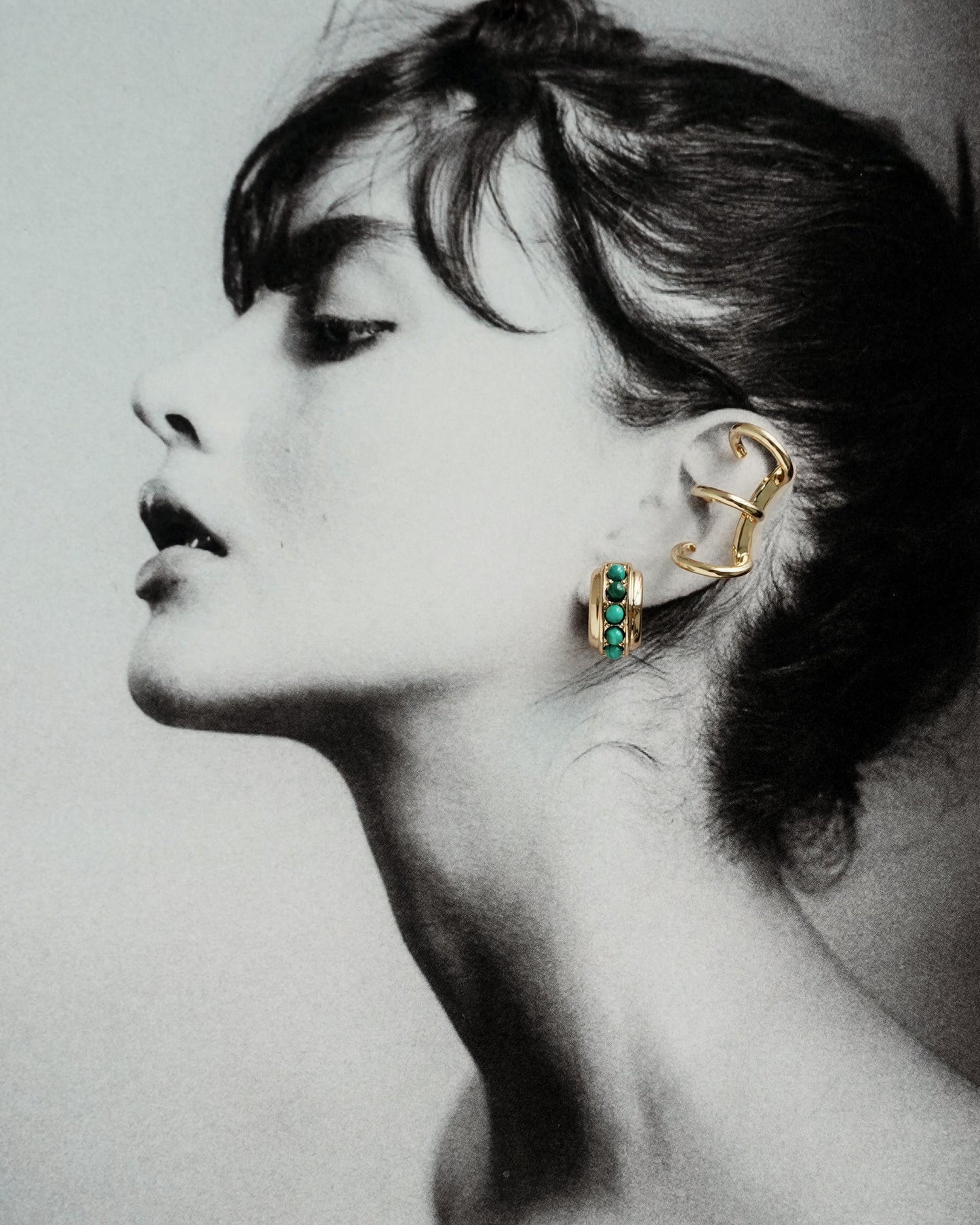 Black and white profile photo of a woman with dark hair, wearing the Mod + Jo Lennon Tube Helix Ear Cuff—two statement, no-piercing gold ear cuffs with green stones and an abstract shape—her head tilted back to showcase them.
