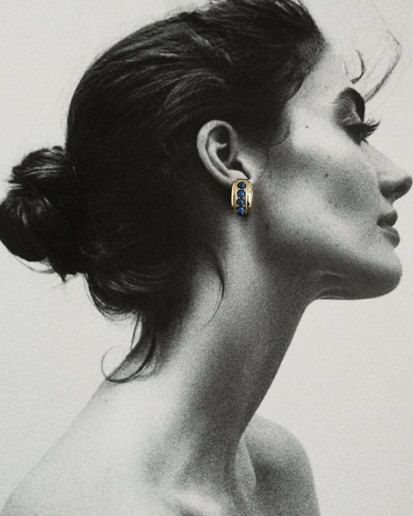Black and white profile of a woman with dark hair in a bun, wearing Mod + Jo's Evy Raised Gemstone Hoops in 18k gold plate set with vibrant blue gemstones; only the hoops are shown in color.