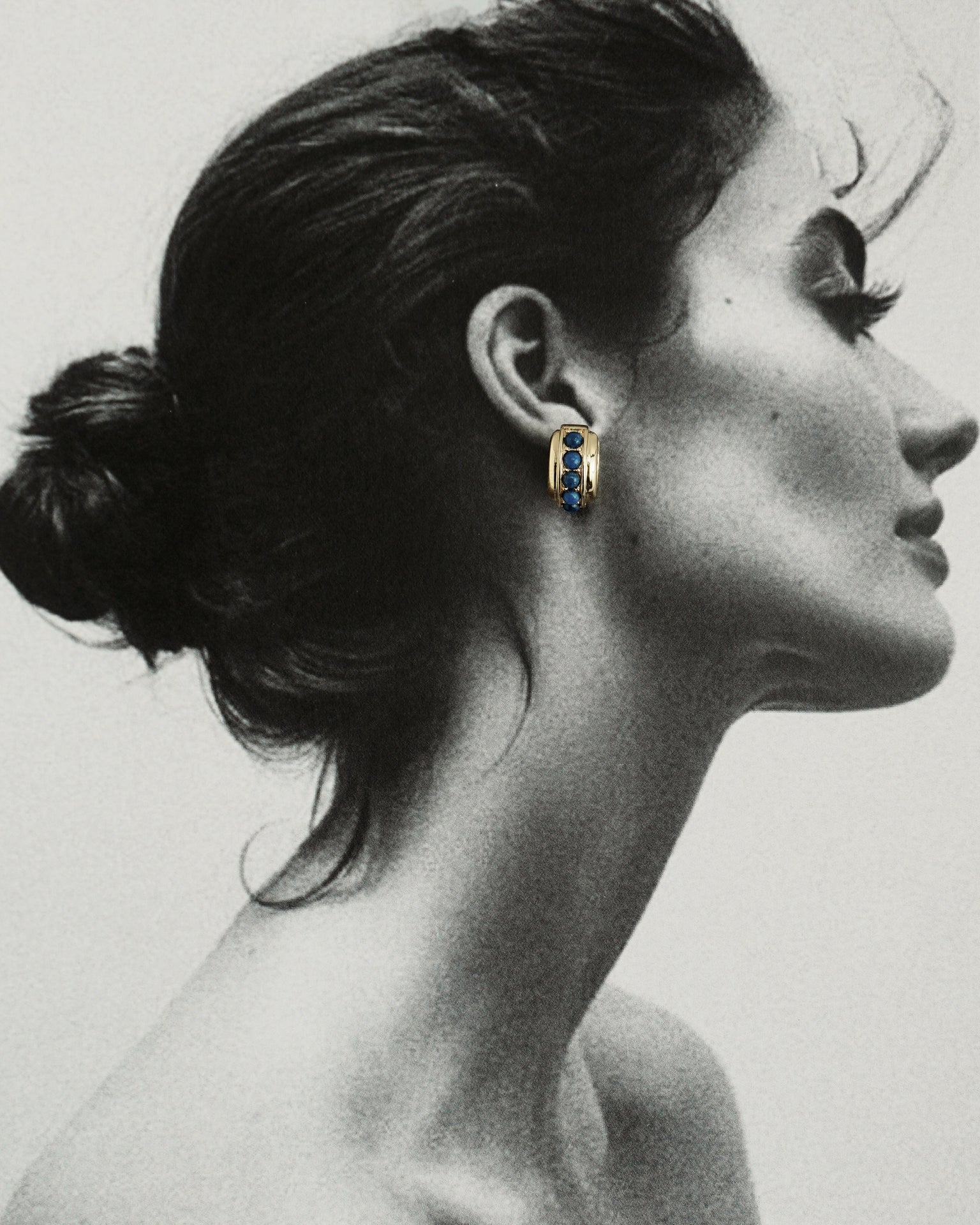 Black and white profile of a woman with dark hair in a bun, wearing Mod + Jo's Evy Raised Gemstone Hoops in 18k gold plate set with vibrant blue gemstones; only the hoops are shown in color.