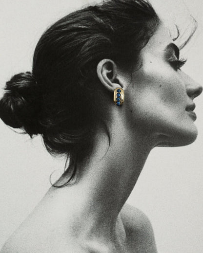 Black and white profile of a woman with dark hair in a bun, wearing Mod + Jo's Evy Raised Gemstone Hoops in 18k gold plate set with vibrant blue gemstones; only the hoops are shown in color.