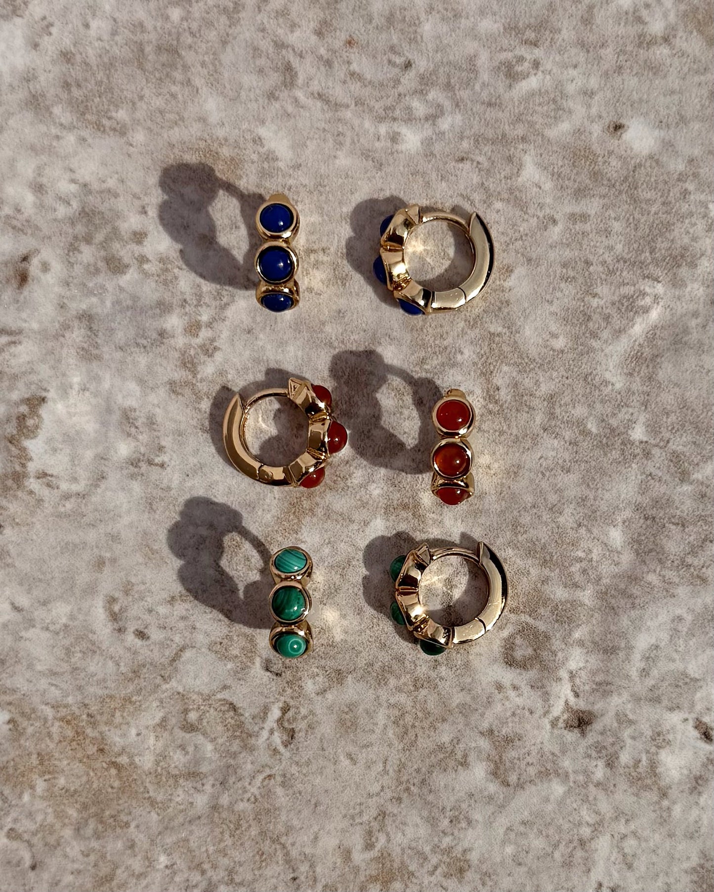 Three pairs of Mod + Jo Corynn Gemstone Huggies with matching studs—blue, red, and genuine malachite green stones—are arranged on a textured stone surface. All are 18k gold plated.