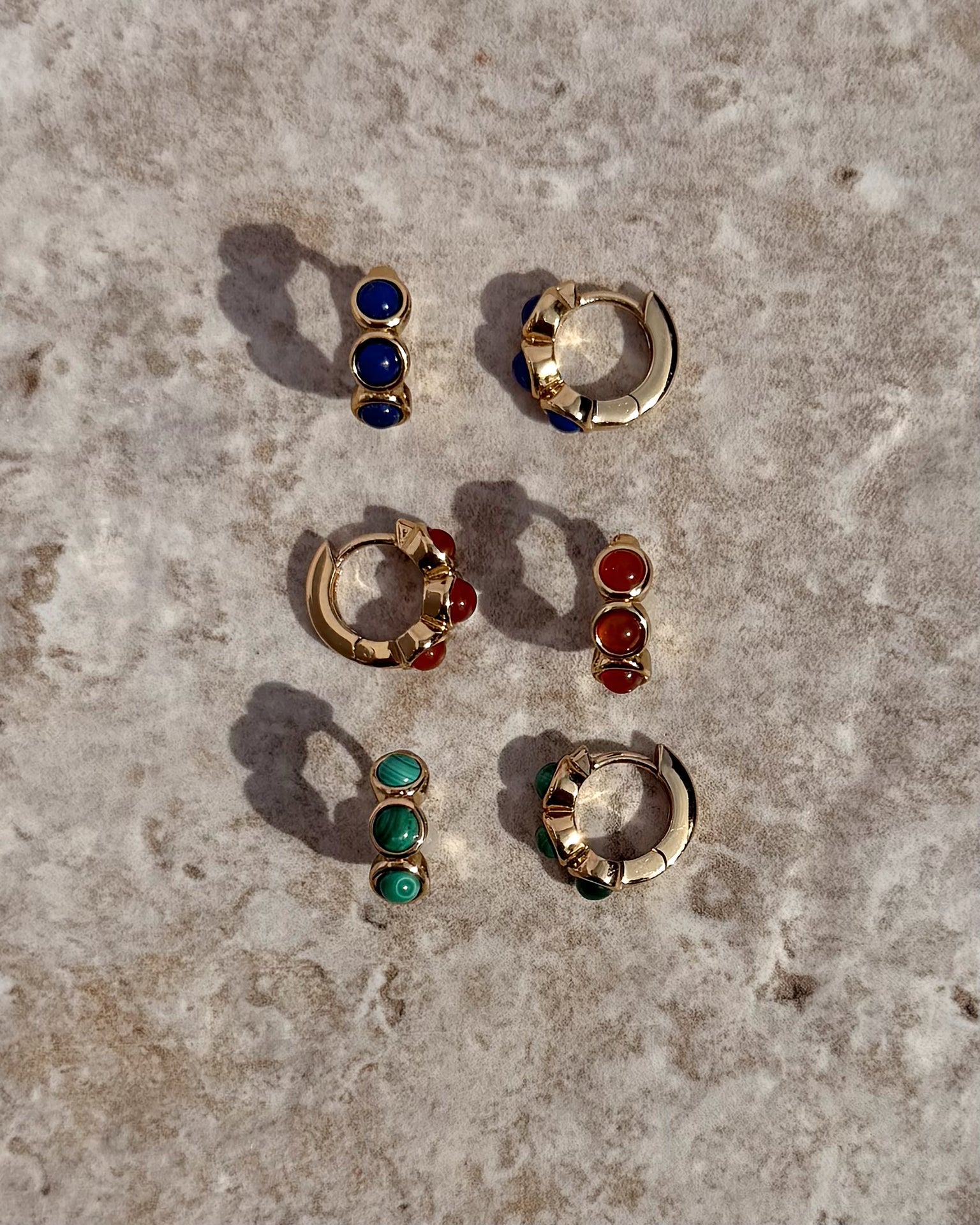 Three pairs of Mod + Jo Corynn Gemstone Huggies with matching studs—blue, red, and genuine malachite green stones—are arranged on a textured stone surface. All are 18k gold plated.