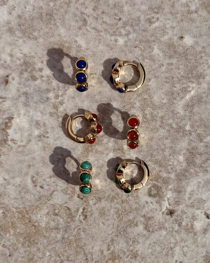 Three pairs of Mod + Jo Corynn Gemstone Huggies with matching studs—blue, red, and genuine malachite green stones—are arranged on a textured stone surface. All are 18k gold plated.