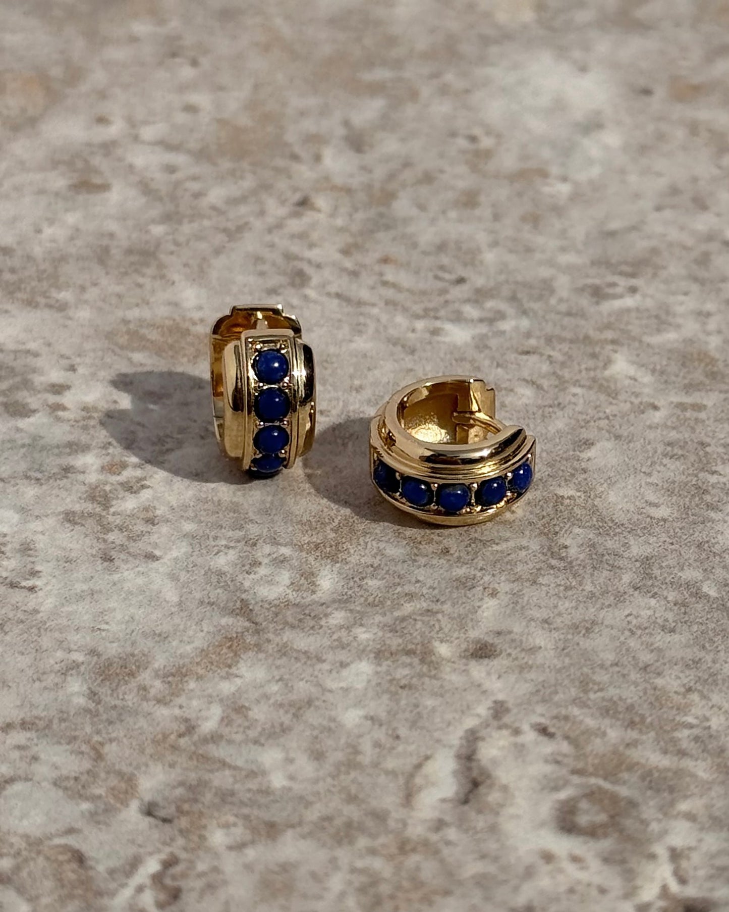 The Evy Raised Gemstone Hoops by Mod + Jo—two small 18k gold-plated hoops, each set with three dark blue stones—are displayed on a textured, light stone surface.