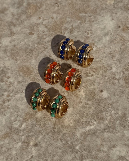Three pairs of Evy Raised Gemstone Hoops by Mod + Jo, crafted with 18k gold plating, are displayed on textured stone. Each pair features a row of blue, red-orange, or genuine malachite green stones for a vibrant touch.