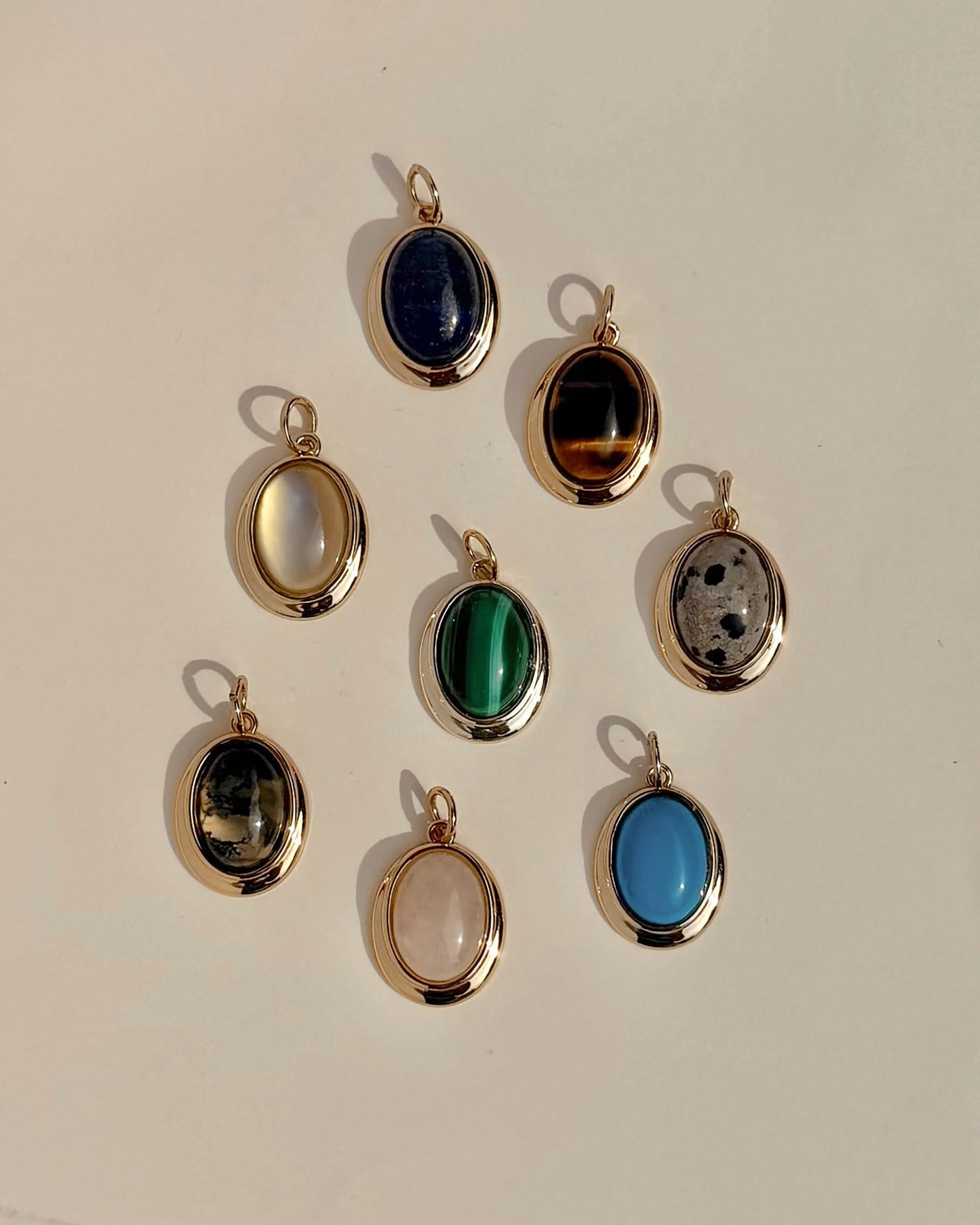 Eight exquisite Rory Gemstone Charms by Mod + Jo, each framed in 14k gold, are elegantly arranged on a light surface. Every charm showcases a distinct gemstone hue—serene blue, vibrant green, mysterious black, luminous white, gentle pink Rose Quartz symbolizing self-love, and uniquely patterned multicolored stones.