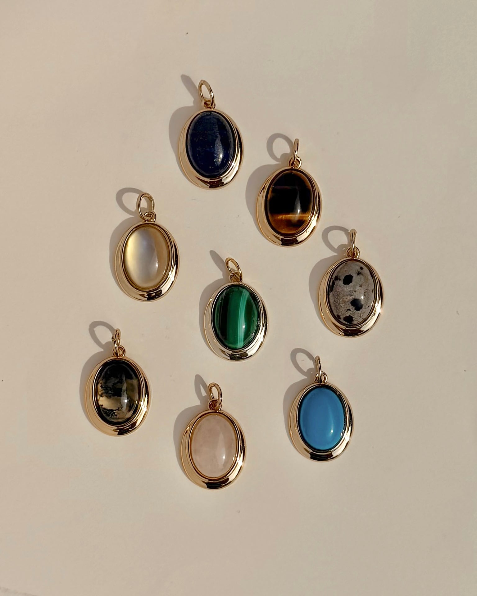 Eight exquisite Rory Gemstone Charms by Mod + Jo, each framed in 14k gold, are elegantly arranged on a light surface. Every charm showcases a distinct gemstone hue—serene blue, vibrant green, mysterious black, luminous white, gentle pink Rose Quartz symbolizing self-love, and uniquely patterned multicolored stones.