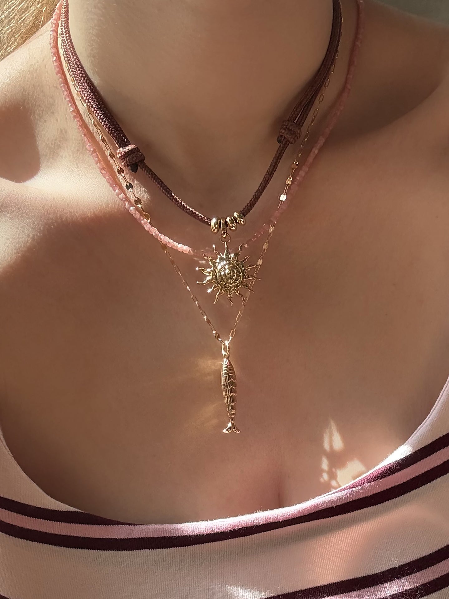A close-up of a person wearing the Mod + Jo Merit Gemstone Necklace, a gold sun pendant necklace, and a pink beaded handmade necklace, styled over a pink-and-white striped top.