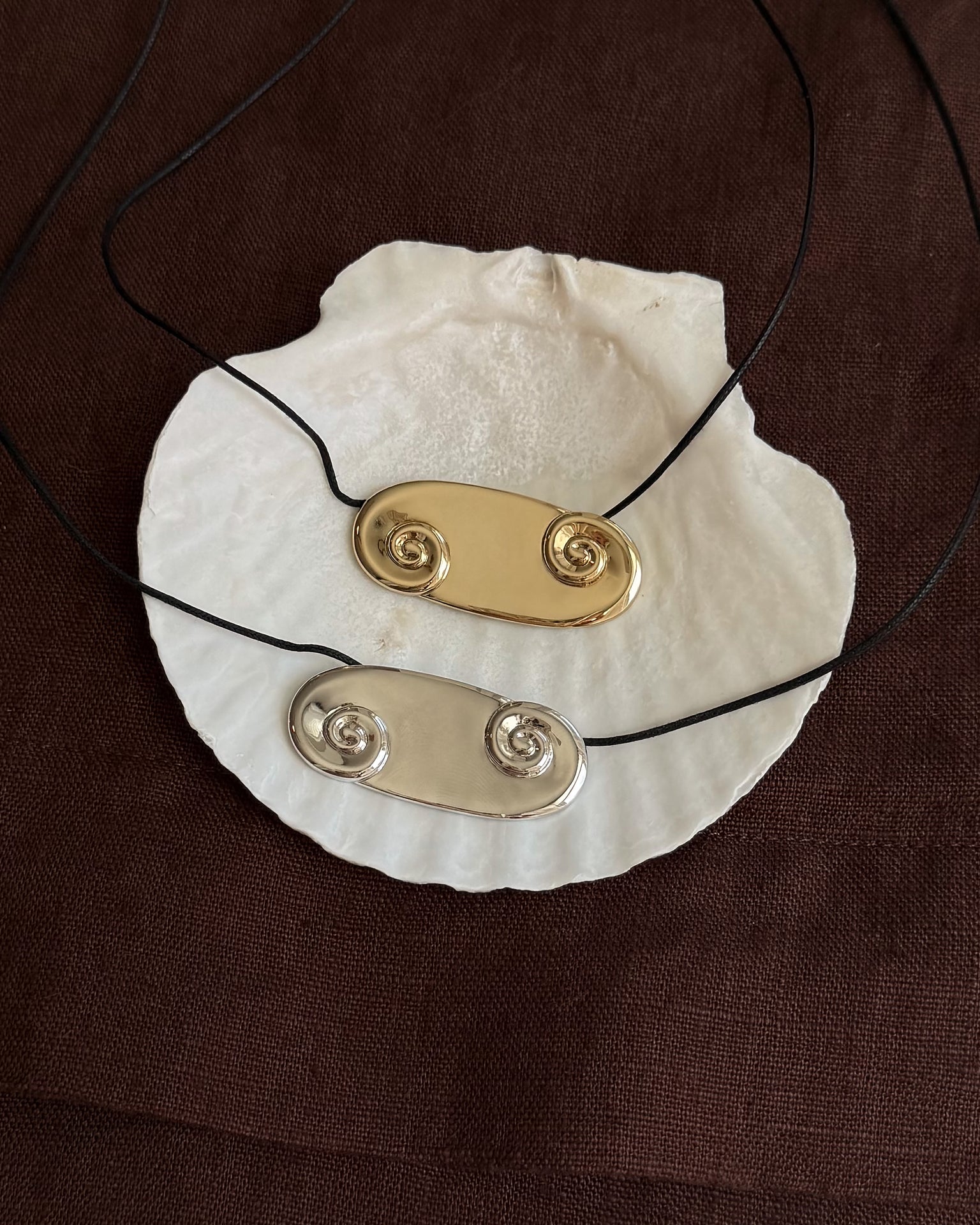 Two Suna Spiral Cord Necklaces by Mod + Jo—one in 14k gold, one in silver—each feature twin spirals on oval pendants. Displayed on a white seashell, both hang from black adjustable bolo cords against brown fabric.