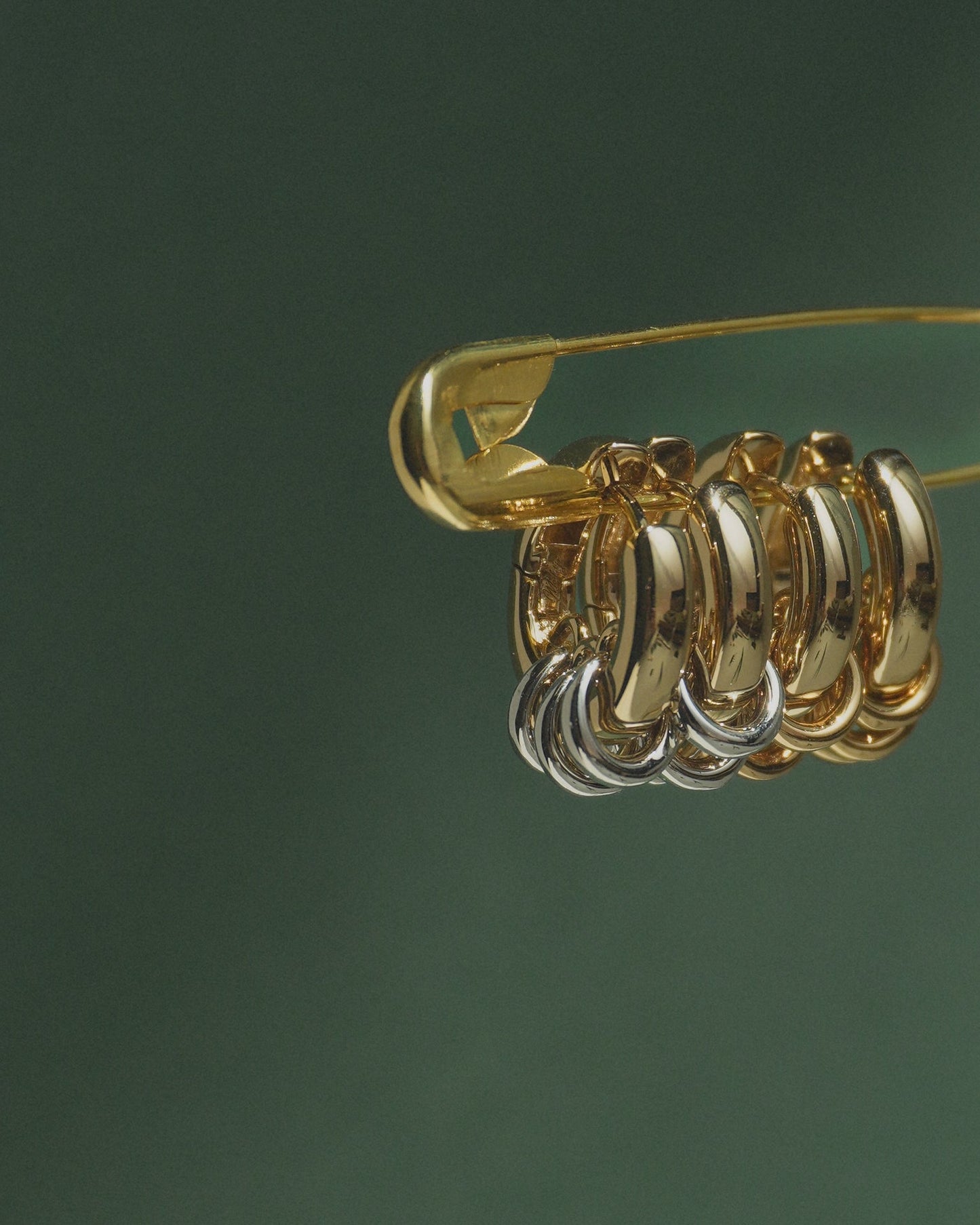 A gold safety pin displays several metallic hoop earrings—primarily gold with a touch of silver—against a dark green background, featuring the Mod + Jo Grande Levi Huggies for an elevated, statement-making style.