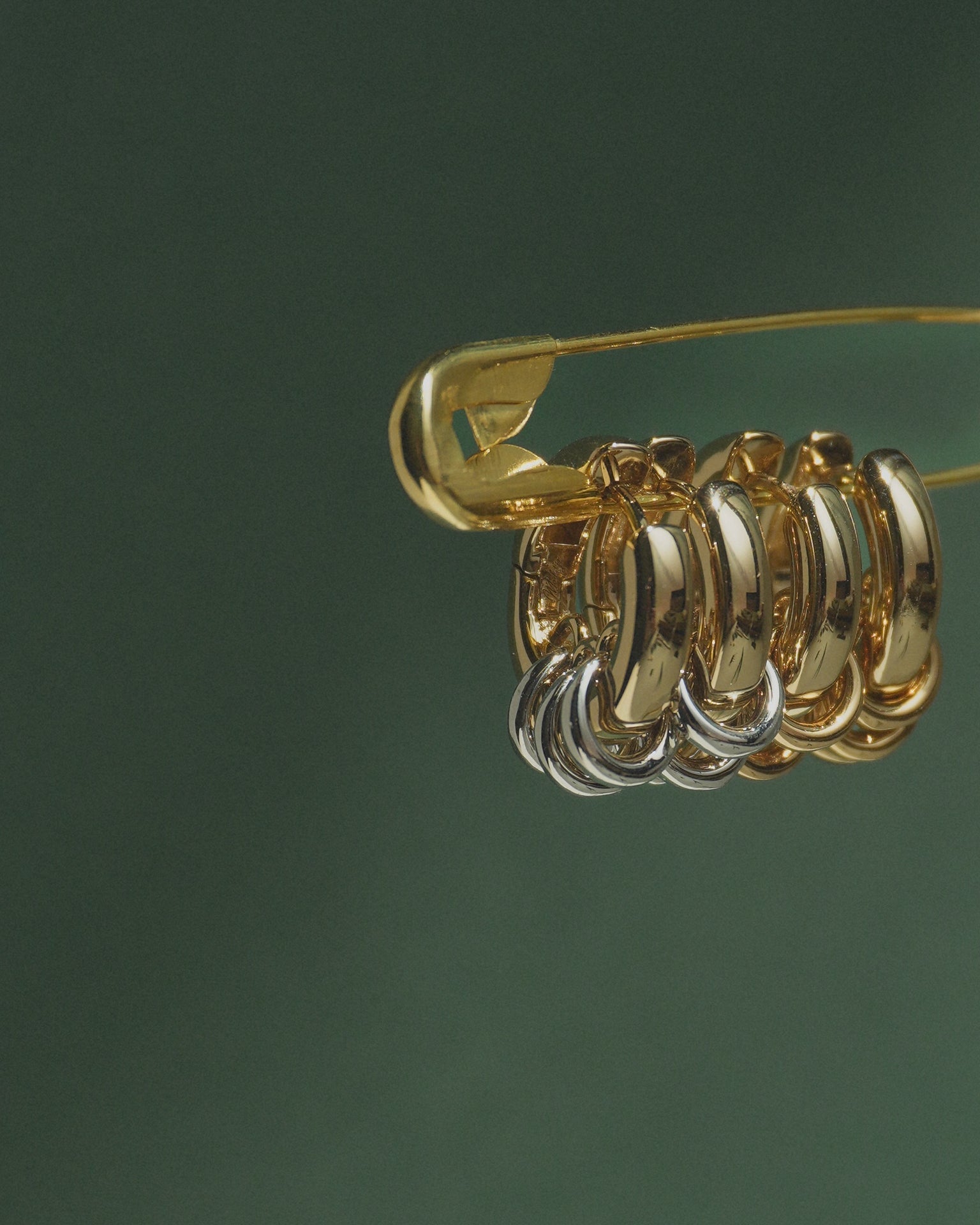 A gold safety pin displays several metallic hoop earrings—primarily gold with a touch of silver—against a dark green background, featuring the Mod + Jo Grande Levi Huggies for an elevated, statement-making style.