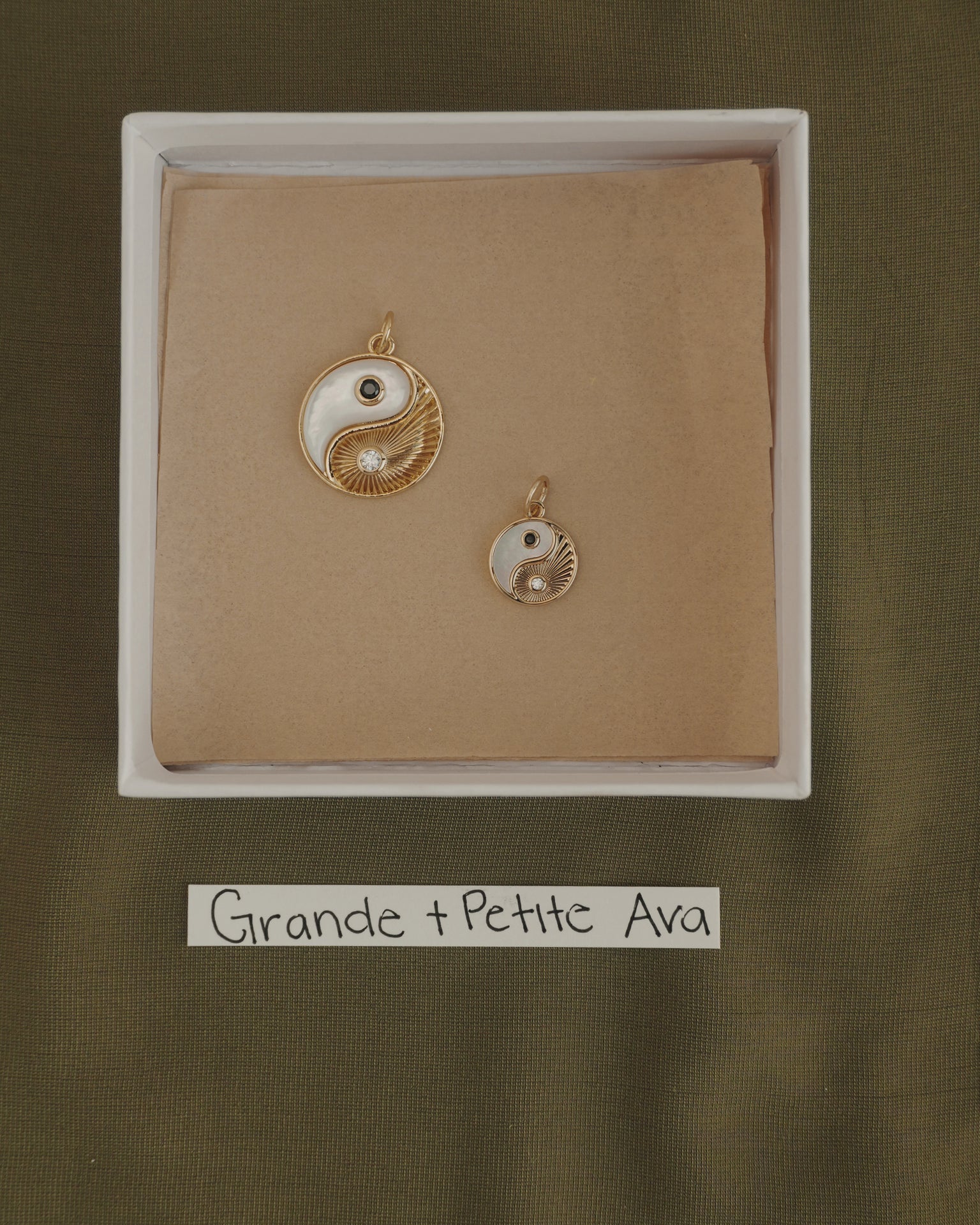Two Mod + Jo Grande Ava Yin Yang charms, one large and one small 14k gold pendant, are displayed in a box on brown paper. Below the box is a label that reads "Grande + Petite Ava" on a green fabric background.
