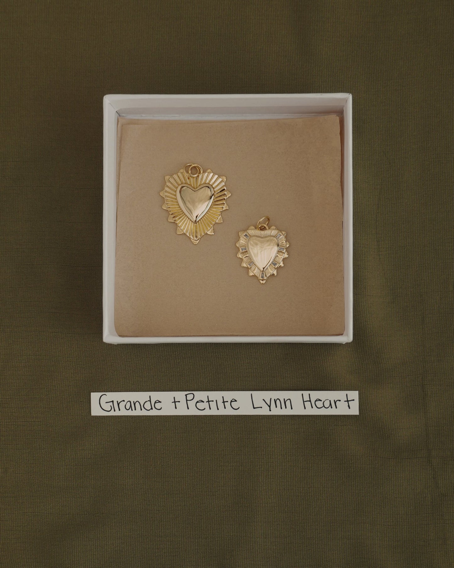 Two gold heart-shaped pendants, one larger and one smaller, are displayed in a box with a brown lining. Each pendant is crafted from 14k gold over brass by Mod + Jo, offering the perfect touch of everyday elegance. A label below reads "Grande + Petite Lynn Heart." The background features olive green fabric. The larger charm is the Grande Lynn Engravable Heart Charm.