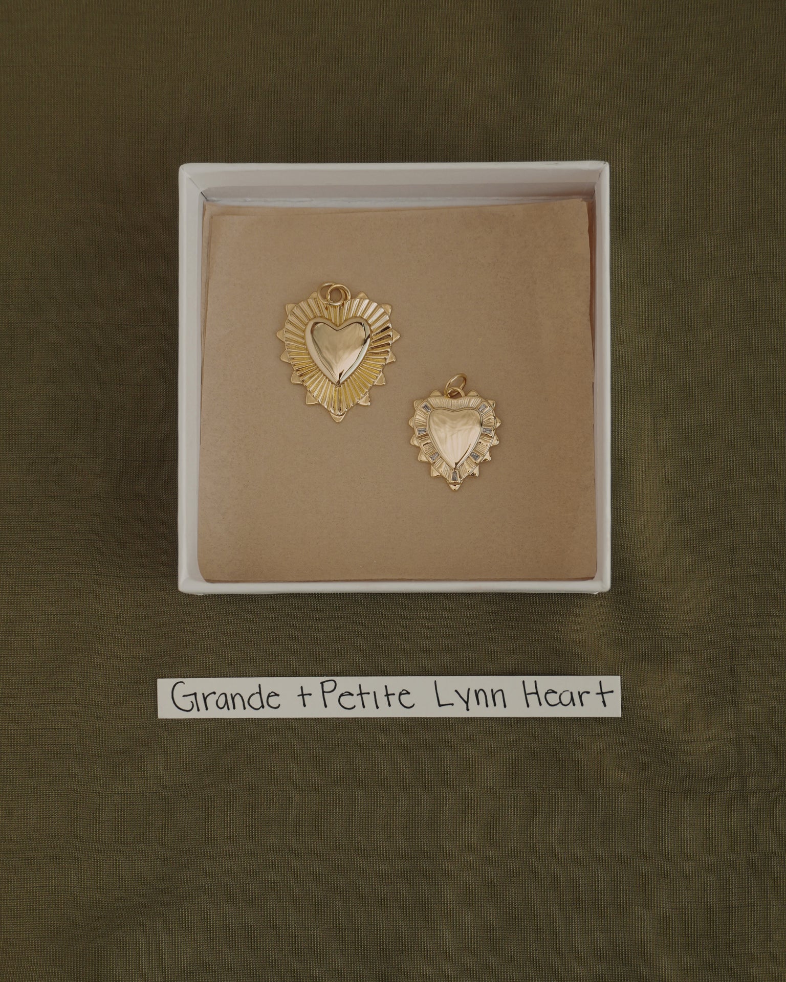 Two gold heart-shaped pendants, one larger and one smaller, are displayed in a box with a brown lining. Each pendant is crafted from 14k gold over brass by Mod + Jo, offering the perfect touch of everyday elegance. A label below reads "Grande + Petite Lynn Heart." The background features olive green fabric. The larger charm is the Grande Lynn Engravable Heart Charm.