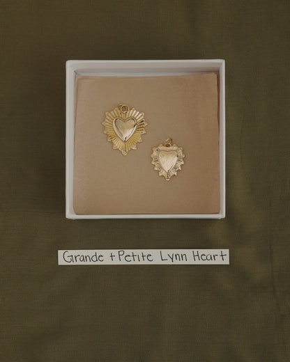 Two gold heart-shaped pendants, one larger and one smaller, are displayed in a box with a brown lining. Each pendant is crafted from 14k gold over brass by Mod + Jo, offering the perfect touch of everyday elegance. A label below reads "Grande + Petite Lynn Heart." The background features olive green fabric. The larger charm is the Grande Lynn Engravable Heart Charm.