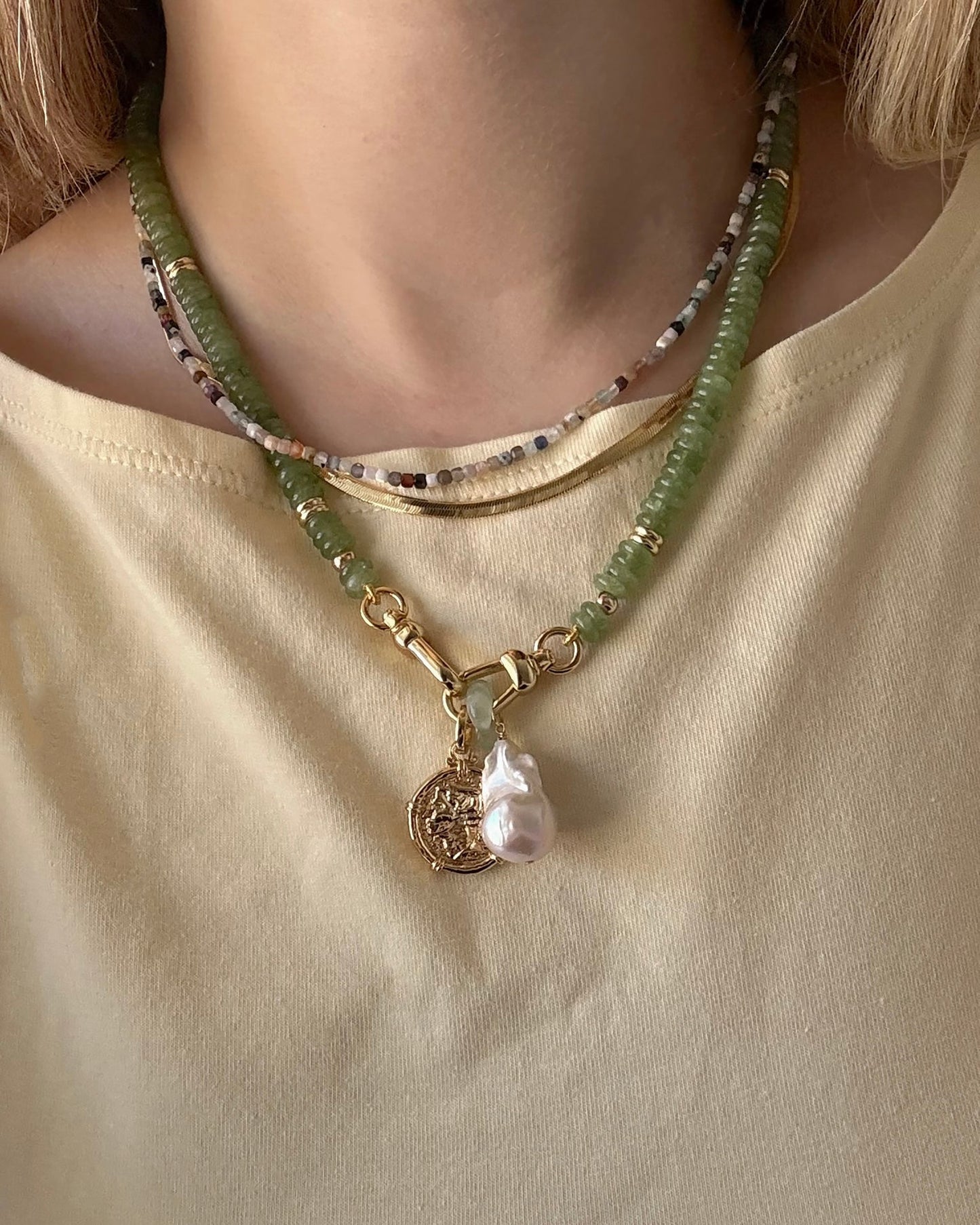 A person in a light yellow shirt wears three layered necklaces: a beaded choker, a multicolored beaded necklace, and Mod + Jo's Jaci Baroque Pearl Charm—a green beaded necklace with 14k gold plated coin and baroque pearl pendant.