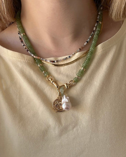 A person in a light yellow shirt wears three layered necklaces: a beaded choker, a multicolored beaded necklace, and Mod + Jo's Jaci Baroque Pearl Charm—a green beaded necklace with 14k gold plated coin and baroque pearl pendant.