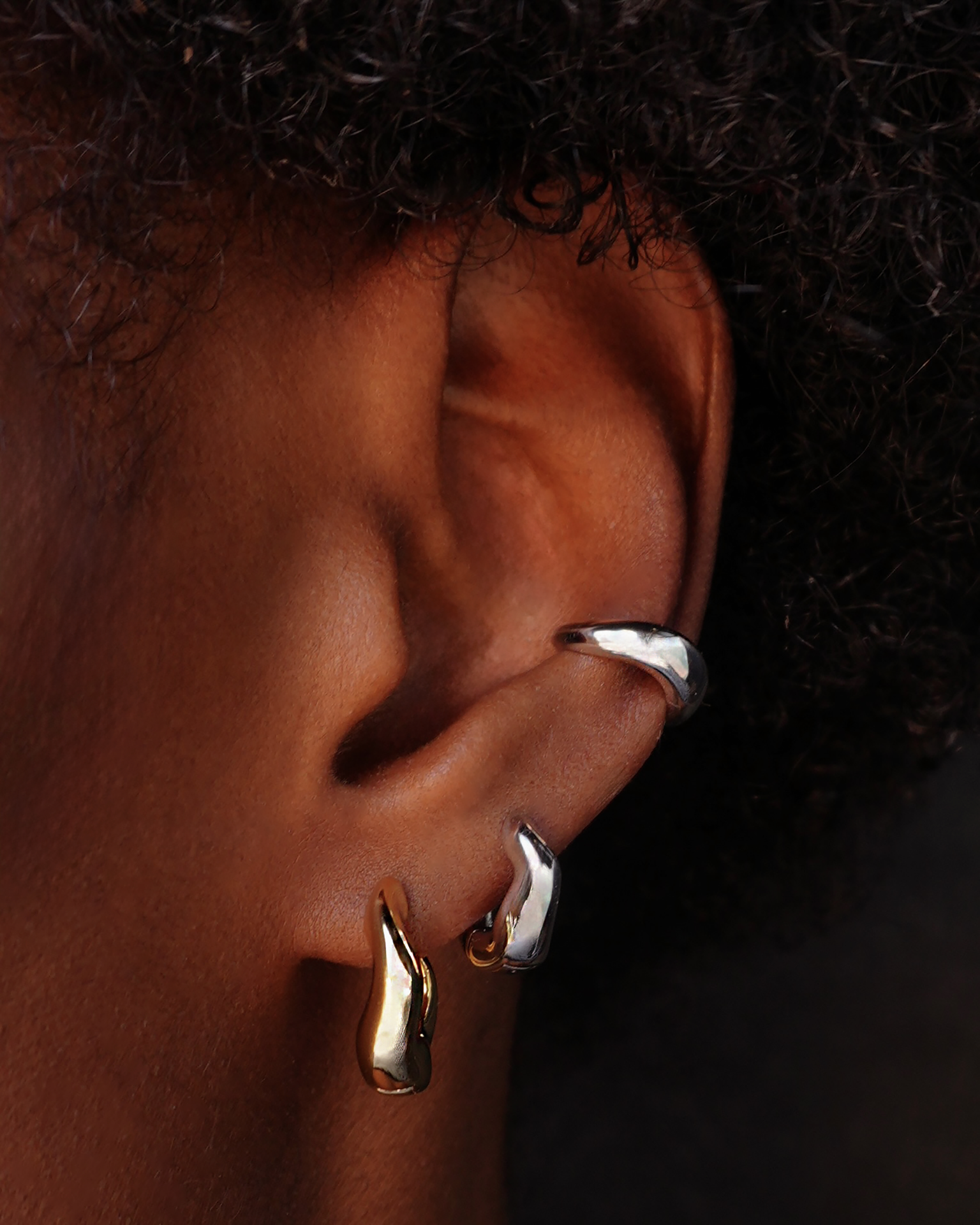 Close-up of an ear wearing gold and silver hoop wavy hoop earrings against dark hair