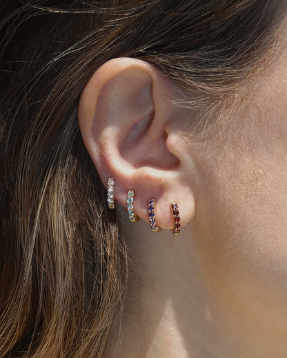 Close-up of a woman's ear wearing four Mod + Jo Harlow Reversible Birthstone Huggies in different gemstone colors, stacked along the lobe. The background is softly blurred.