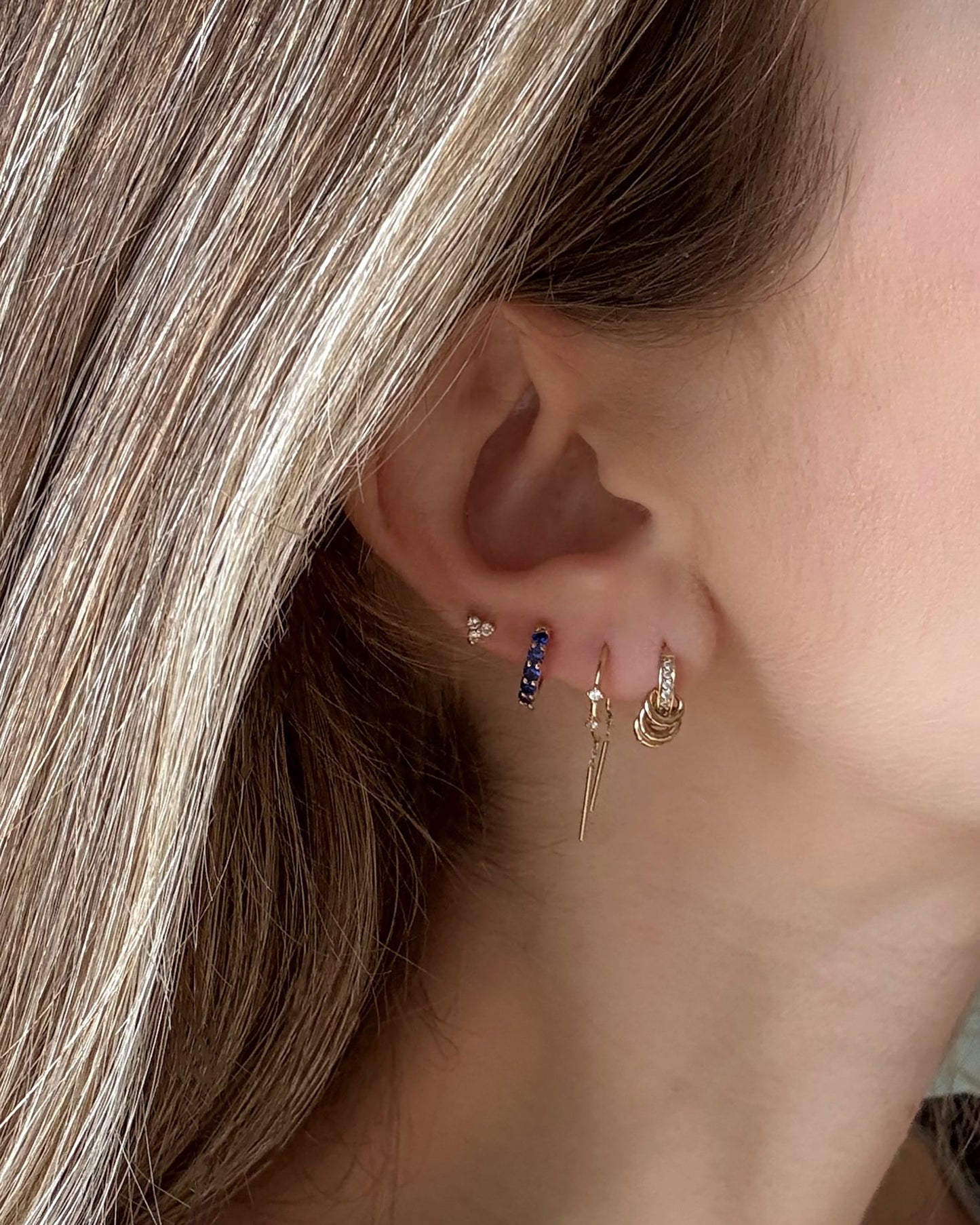 Close-up of a woman's ear with multiple piercings, showcasing Mod + Jo's Harlow Reversible Birthstone Huggies, 18k gold plated with gemstones, alongside other hoops and studs. Light brown hair partially visible.