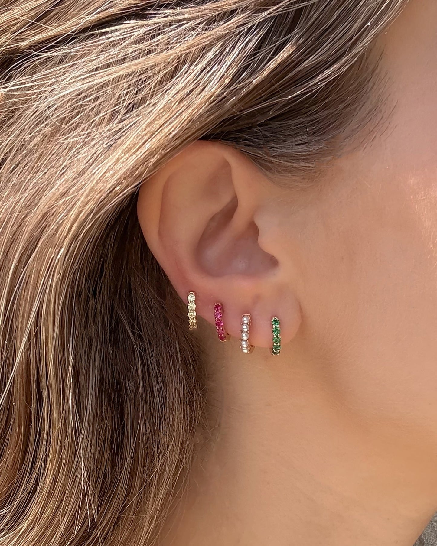 Close-up of a woman’s ear with four Harlow Reversible Birthstone Huggies by Mod + Jo in gold, pink, white, and green 18k gold plating with matching gemstones—her light brown hair tucked back to highlight these colorful earrings.