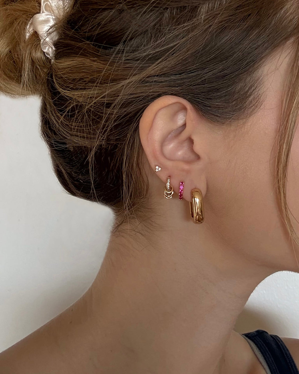 A close-up of a woman's ear wearing the Mod + Jo Harlow Reversible Birthstone Huggies. Her hair is tied back with a white scrunchie, and she is dressed in a dark top.