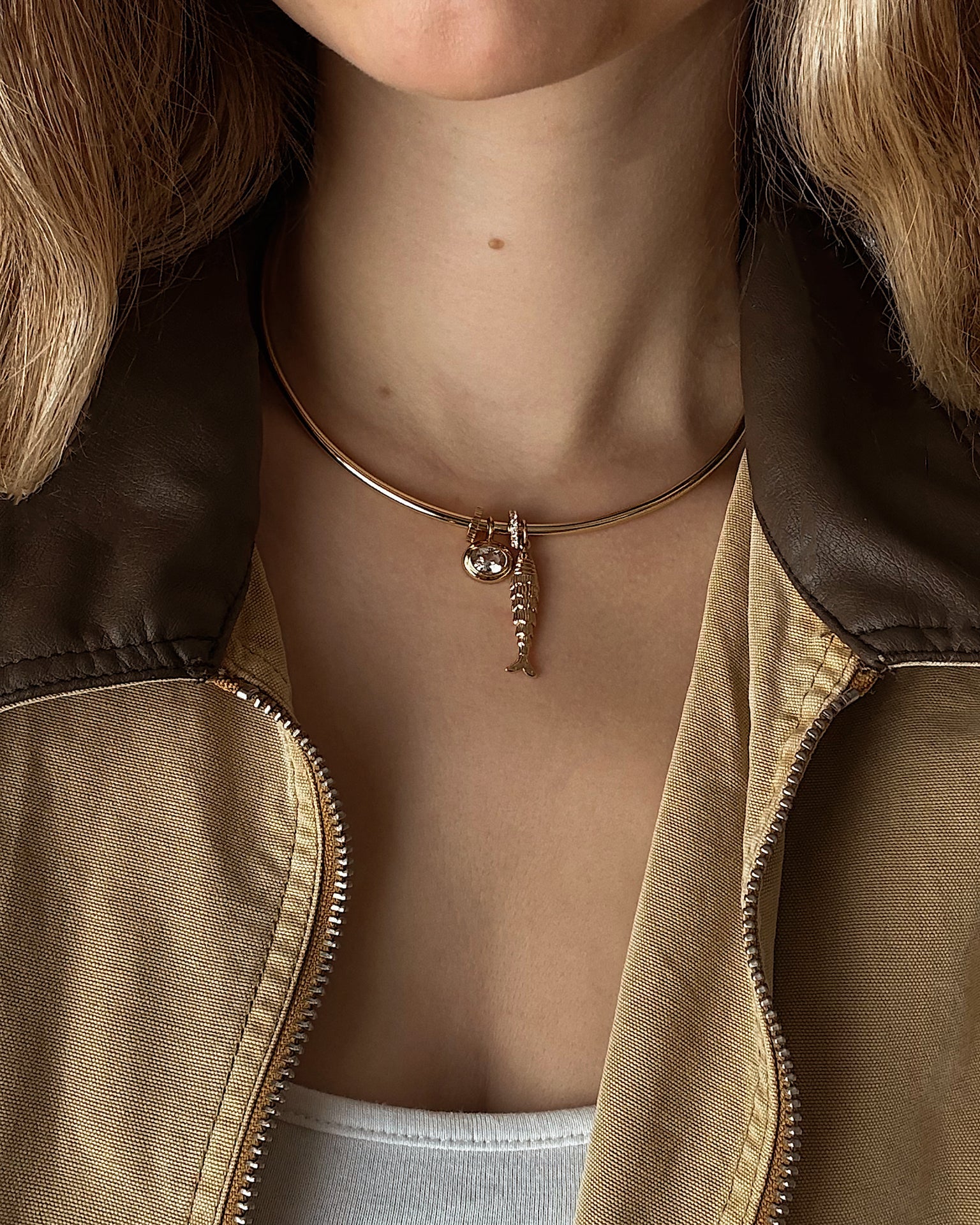 A woman in a brown and tan jacket with a white top wears a gold choker necklace featuring two pendants: a small fish skeleton and the Mod + Jo Marla Sapphire Bezel Charm.
