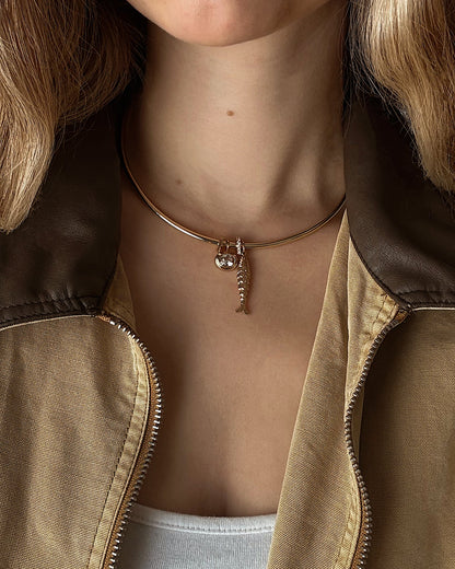 A woman in a brown and tan jacket with a white top wears a gold choker necklace featuring two pendants: a small fish skeleton and the Mod + Jo Marla Sapphire Bezel Charm.
