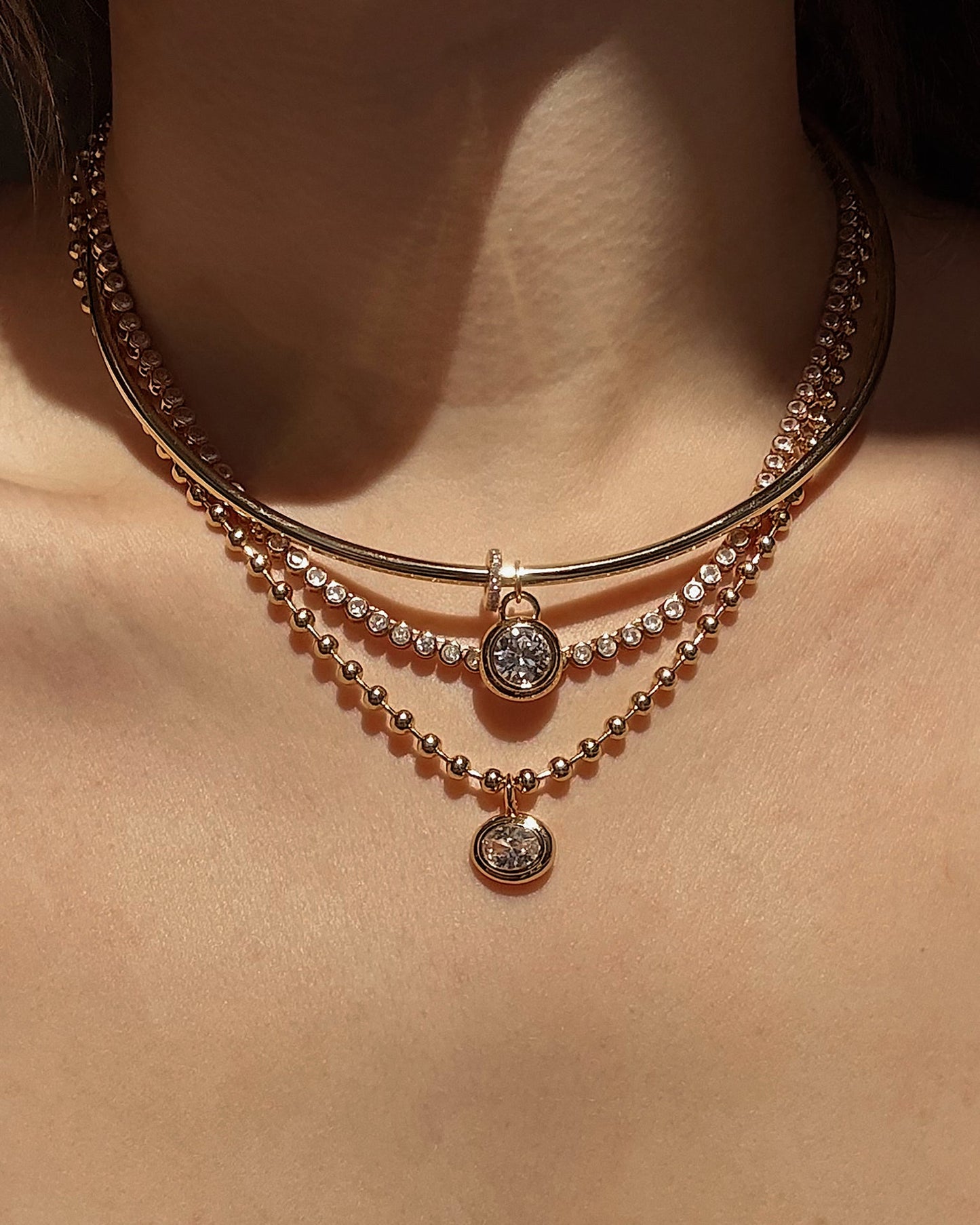 A close-up of someone wearing three layered gold necklaces, featuring Mod + Jo's Marla Sapphire Bezel Charm with lab-grown gemstones and a delicate 14k gold pendant, against bare skin in warm lighting.