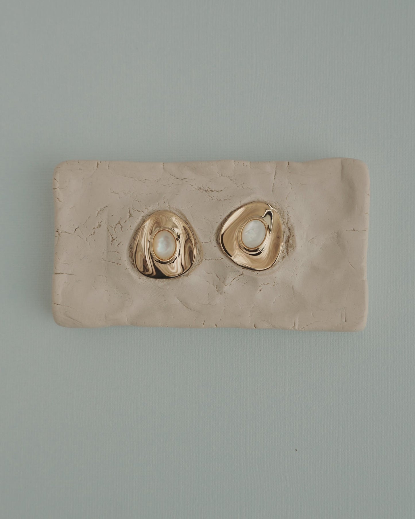 The Mod + Jo Hunter Statement Studs, featuring 14k gold-toned irregular shapes with pearl centers, are shown on a textured beige clay slab, set against a light blue background.