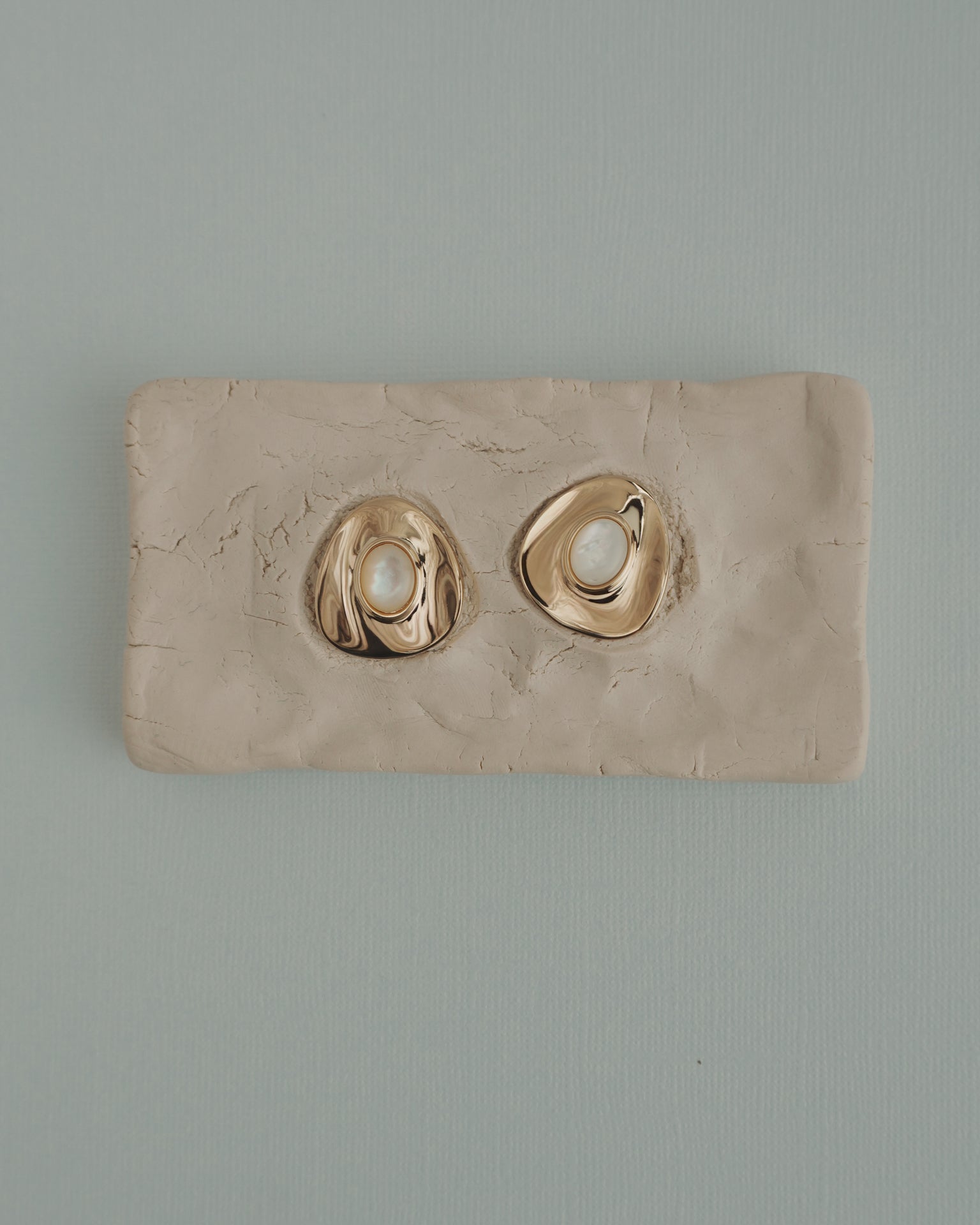 The Mod + Jo Hunter Statement Studs, featuring 14k gold-toned irregular shapes with pearl centers, are shown on a textured beige clay slab, set against a light blue background.