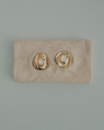 The Mod + Jo Hunter Statement Studs, featuring 14k gold-toned irregular shapes with pearl centers, are shown on a textured beige clay slab, set against a light blue background.