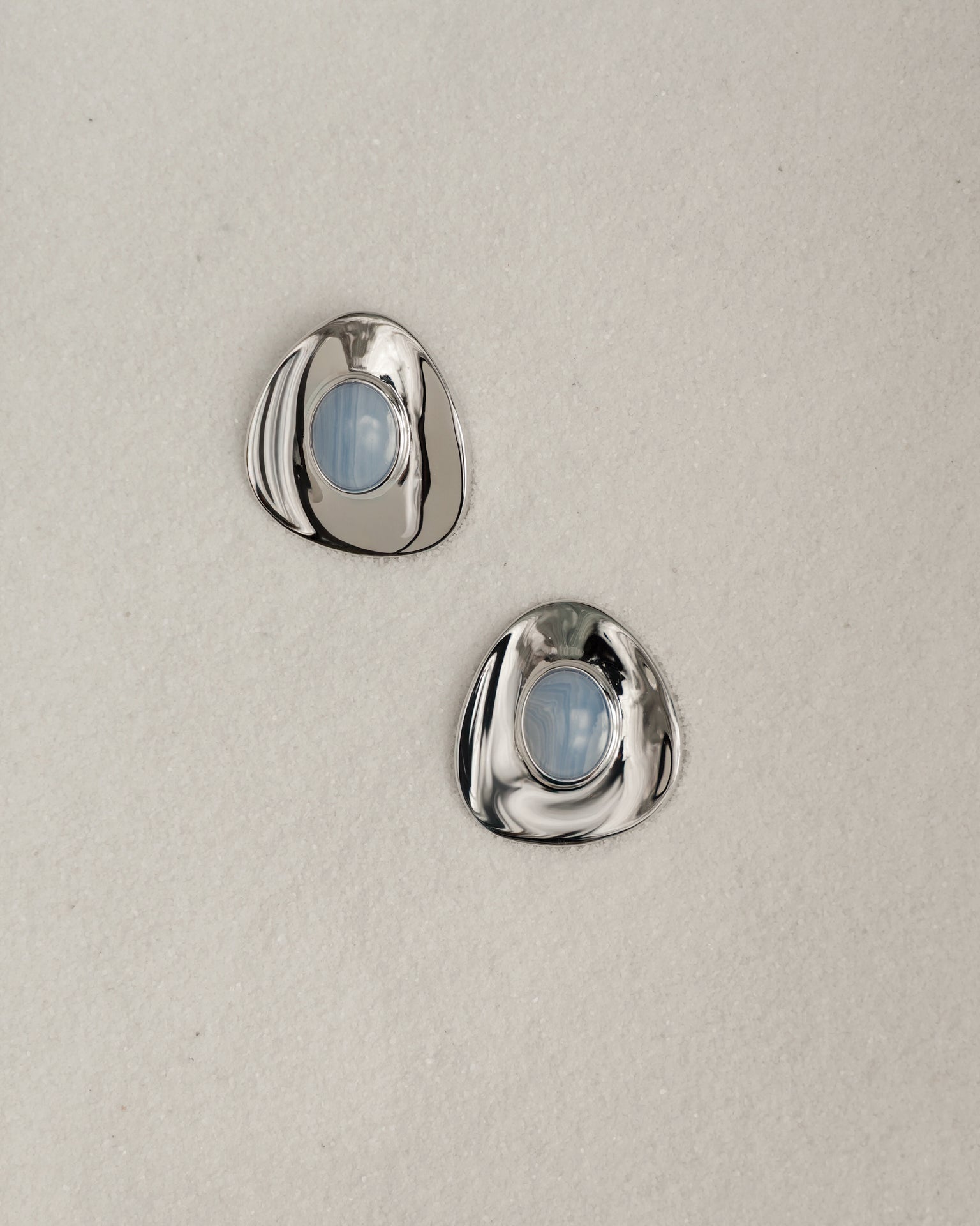 The Mod + Jo Hunter Statement Studs are two shiny, irregular silver earrings with round blue chalcedony centers, displayed on a light beige textured surface.