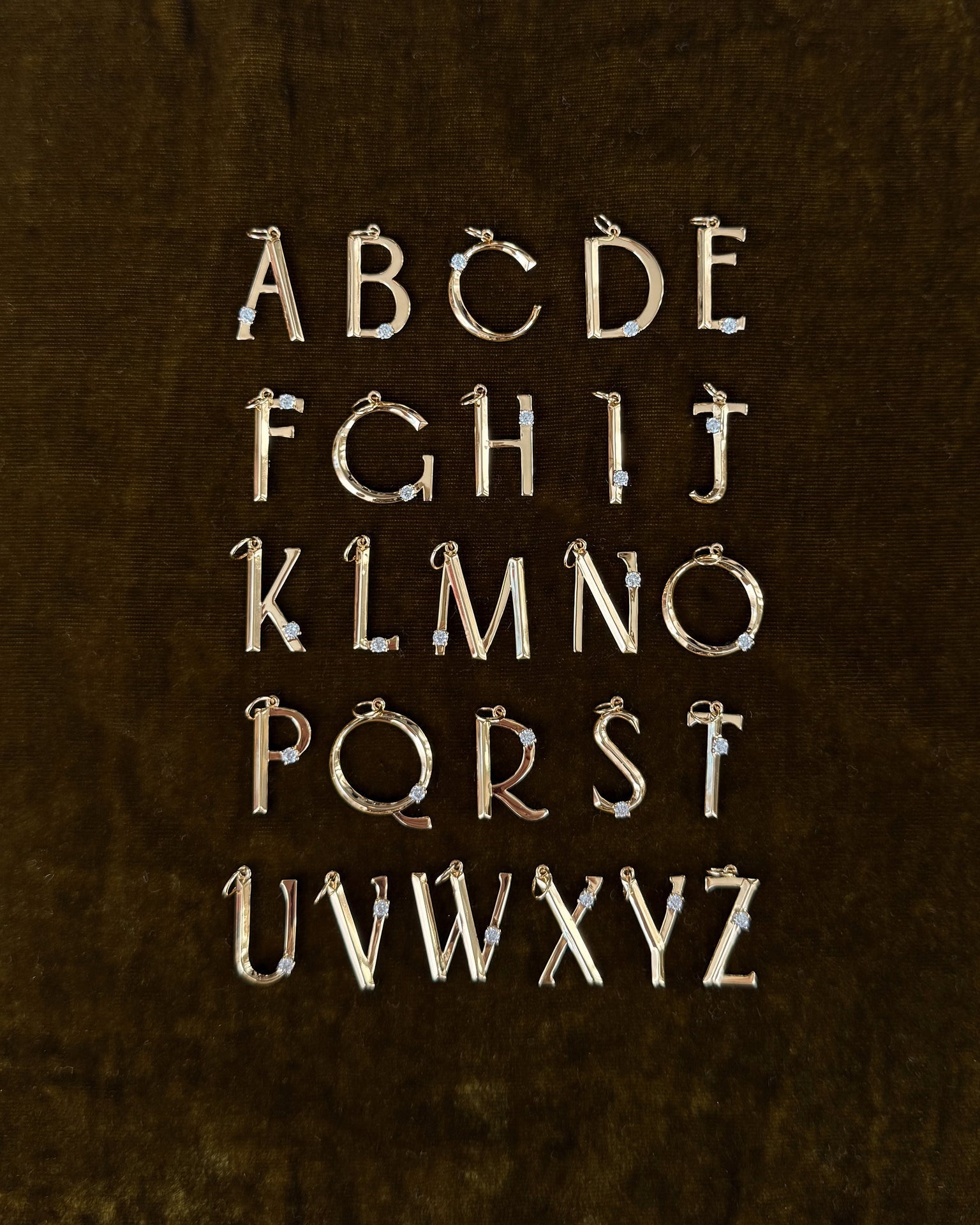 The Eloise Letter Charm by Mod + Jo features gold uppercase alphabet pendants with sparkling cubic zirconia accents, arranged in five rows on a dark brown velvet background, displaying A to Z.