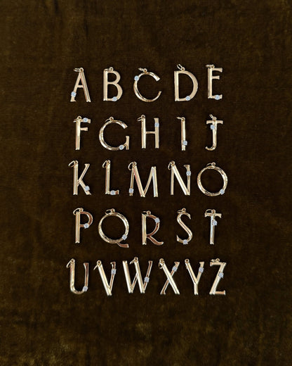 The Eloise Letter Charm by Mod + Jo features gold uppercase alphabet pendants with sparkling cubic zirconia accents, arranged in five rows on a dark brown velvet background, displaying A to Z.