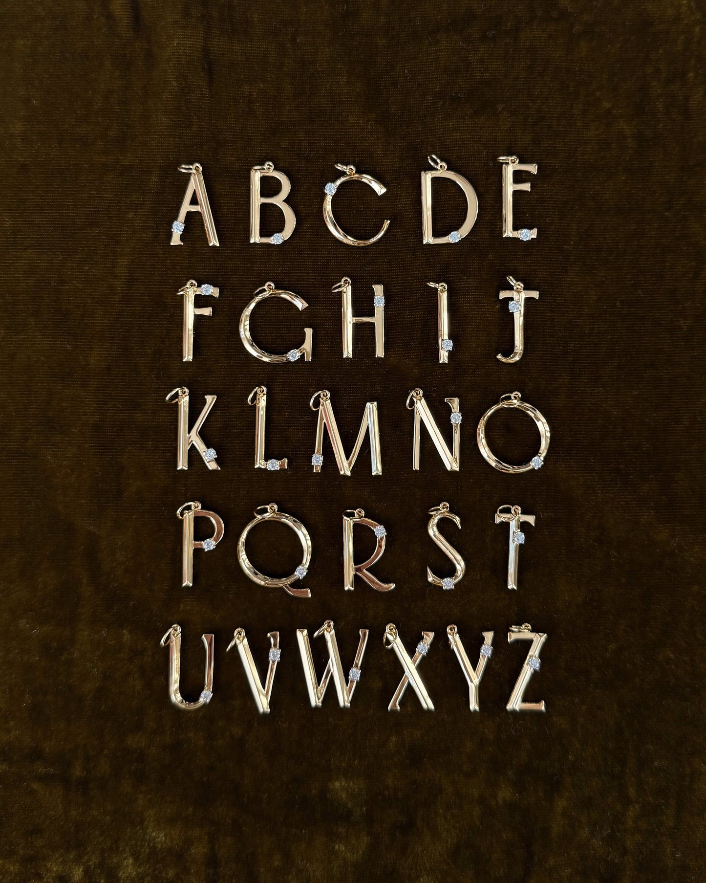 The Eloise Letter Charm by Mod + Jo features gold uppercase alphabet pendants with sparkling cubic zirconia accents, arranged in five rows on a dark brown velvet background, displaying A to Z.