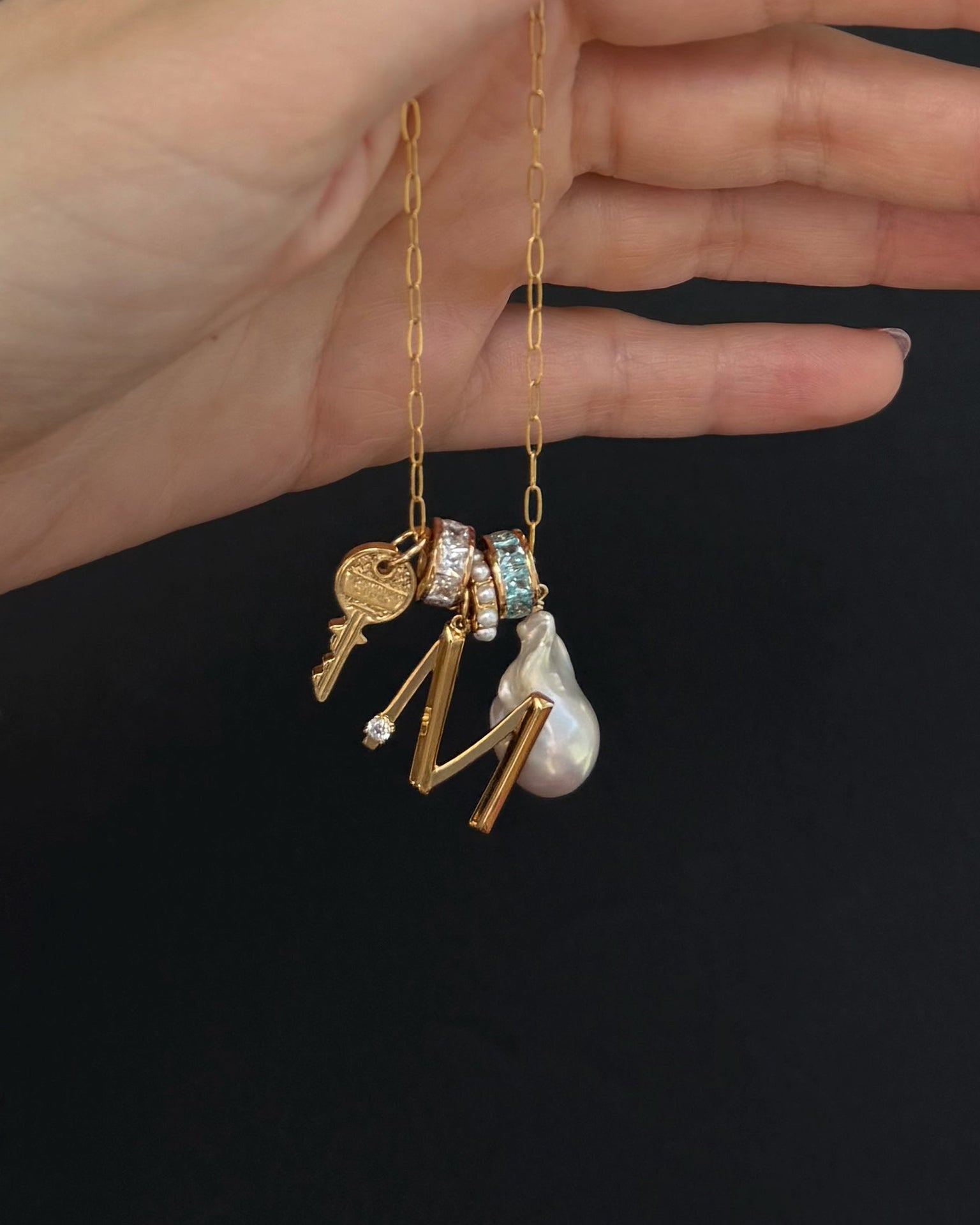A hand holds the Mod + Jo Alden Pearl Charm Spacer, a tarnish-resistant gold chain necklace featuring charms like a key, two crystal-studded rings, an M, pearl drop, shell pearls, and a small round pendant against a black background.