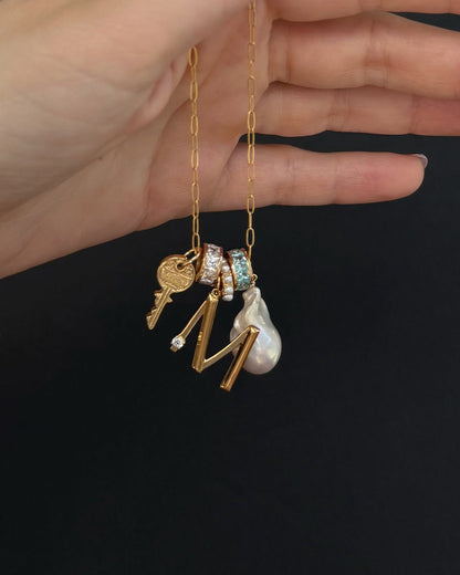 A hand holds the Mod + Jo Alden Pearl Charm Spacer, a tarnish-resistant gold chain necklace featuring charms like a key, two crystal-studded rings, an M, pearl drop, shell pearls, and a small round pendant against a black background.
