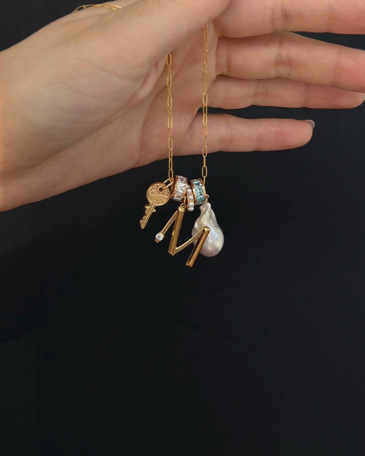A hand holds Mod + Jo’s Frankie Key Charm gold chain necklace with charms—a key, two gemstone rings, a teardrop pearl, and a gold letter M—against a black background.