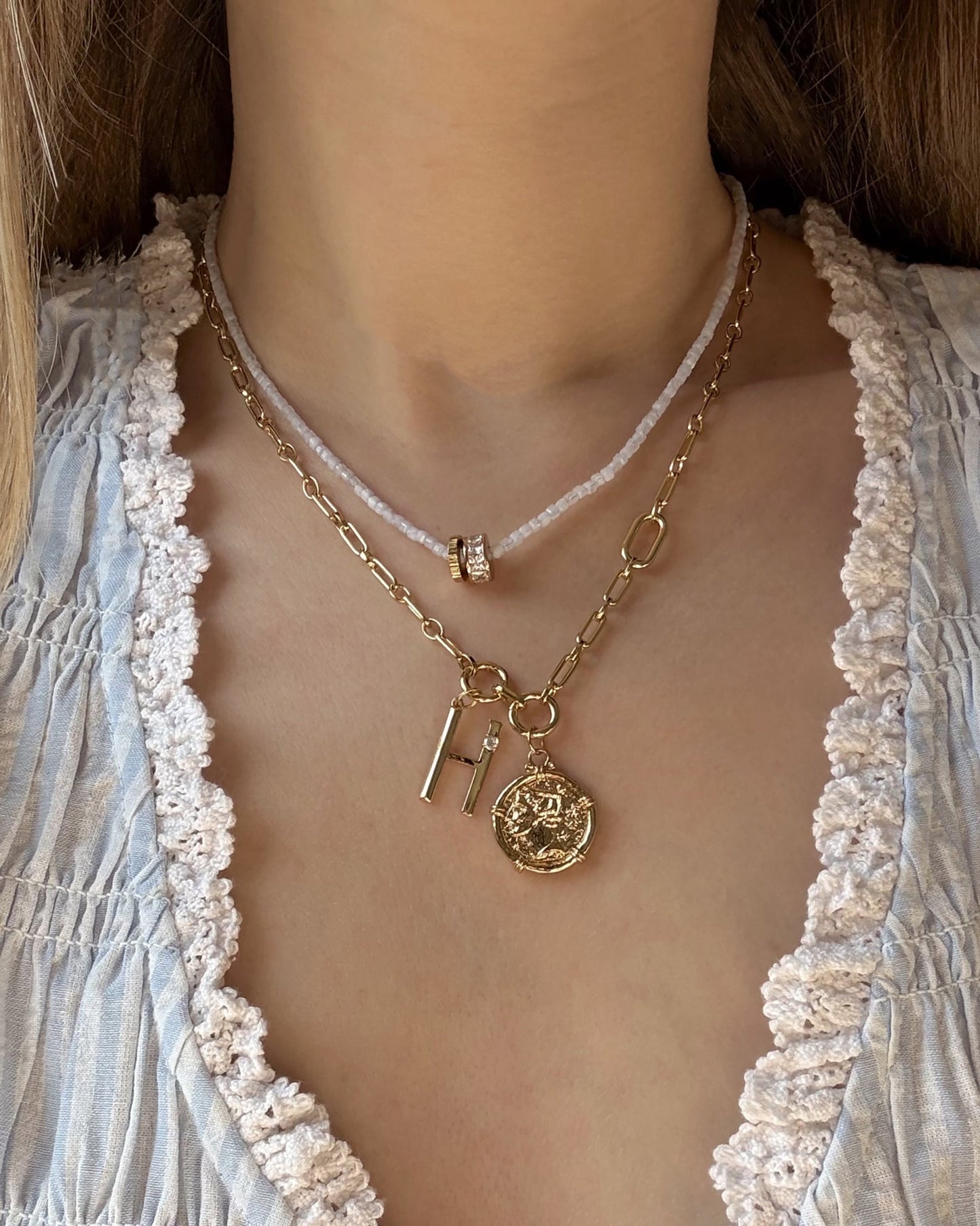 A close-up of a woman's neck wearing three layered necklaces, including Mod + Jo’s Este Birthstone Charm Spacer, with a thin white cord and gold ring, coin pendant with initials, paired with a light blue textured blouse.
