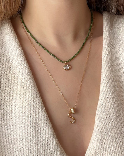 A person wears a cream knit top and two layered necklaces: the Mod + Jo Merit Gemstone Necklace and a thin gold chain with an “S” pendant and small crystal charm.