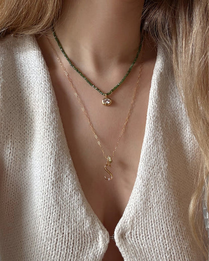 A woman wears a cream knit top and two layered necklaces: the Mod + Jo Este Birthstone Charm Spacer with green beads, and a gold chain with a gold letter S—showcasing her style and love for personalized jewelry.
